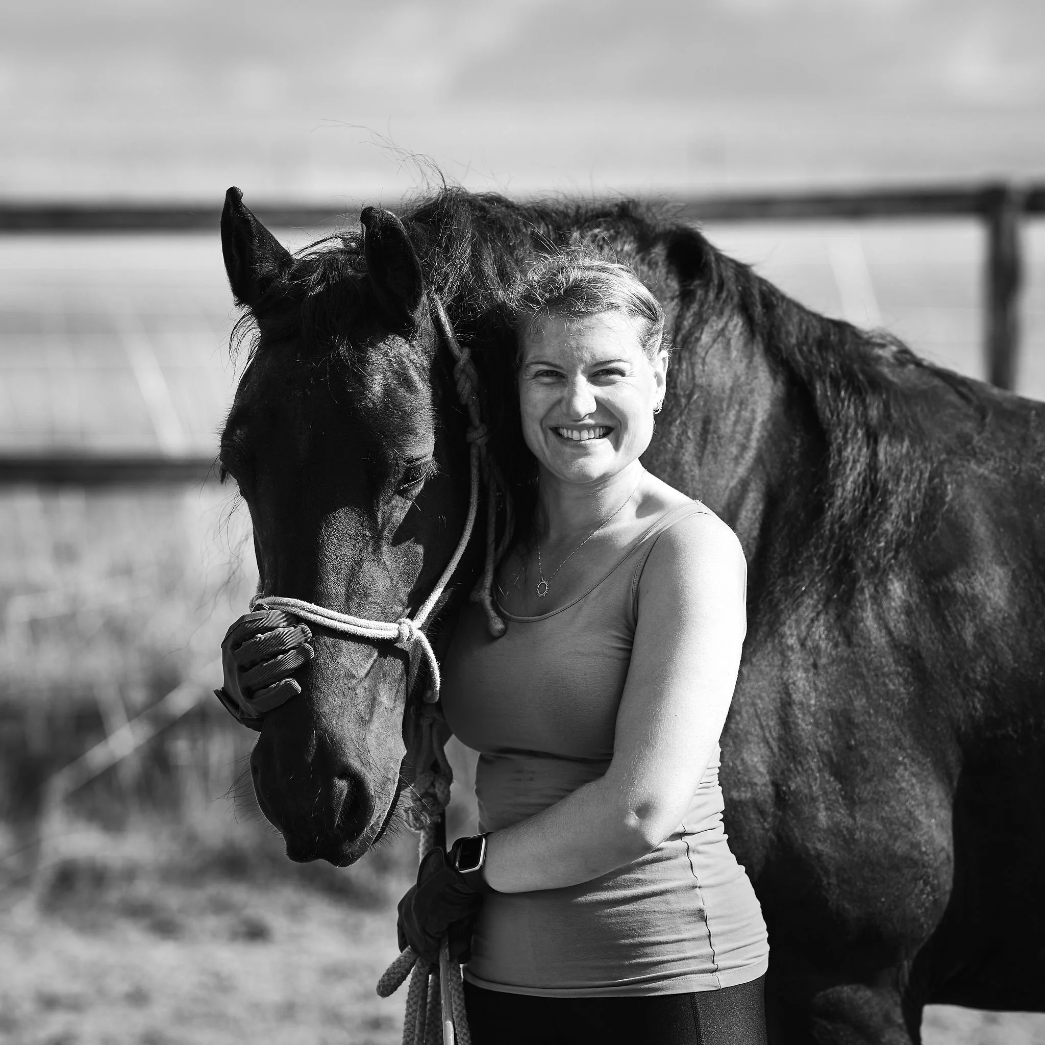 A woman smiling while holding a horse by its bridle outdoors on a cloudy day, with a fence and open field in the background.