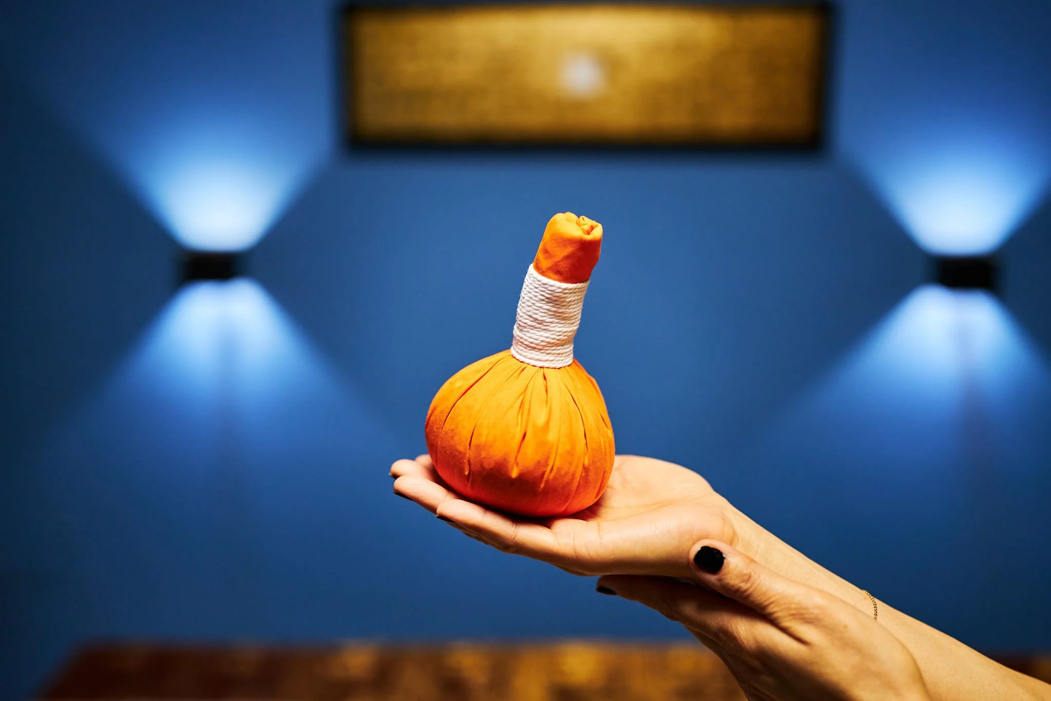 A person's hand holding a small orange stuffed pumpkin with a carrot nose and a white string wrapped around its stem, set against a blue background with a television screen or board above in the distance.