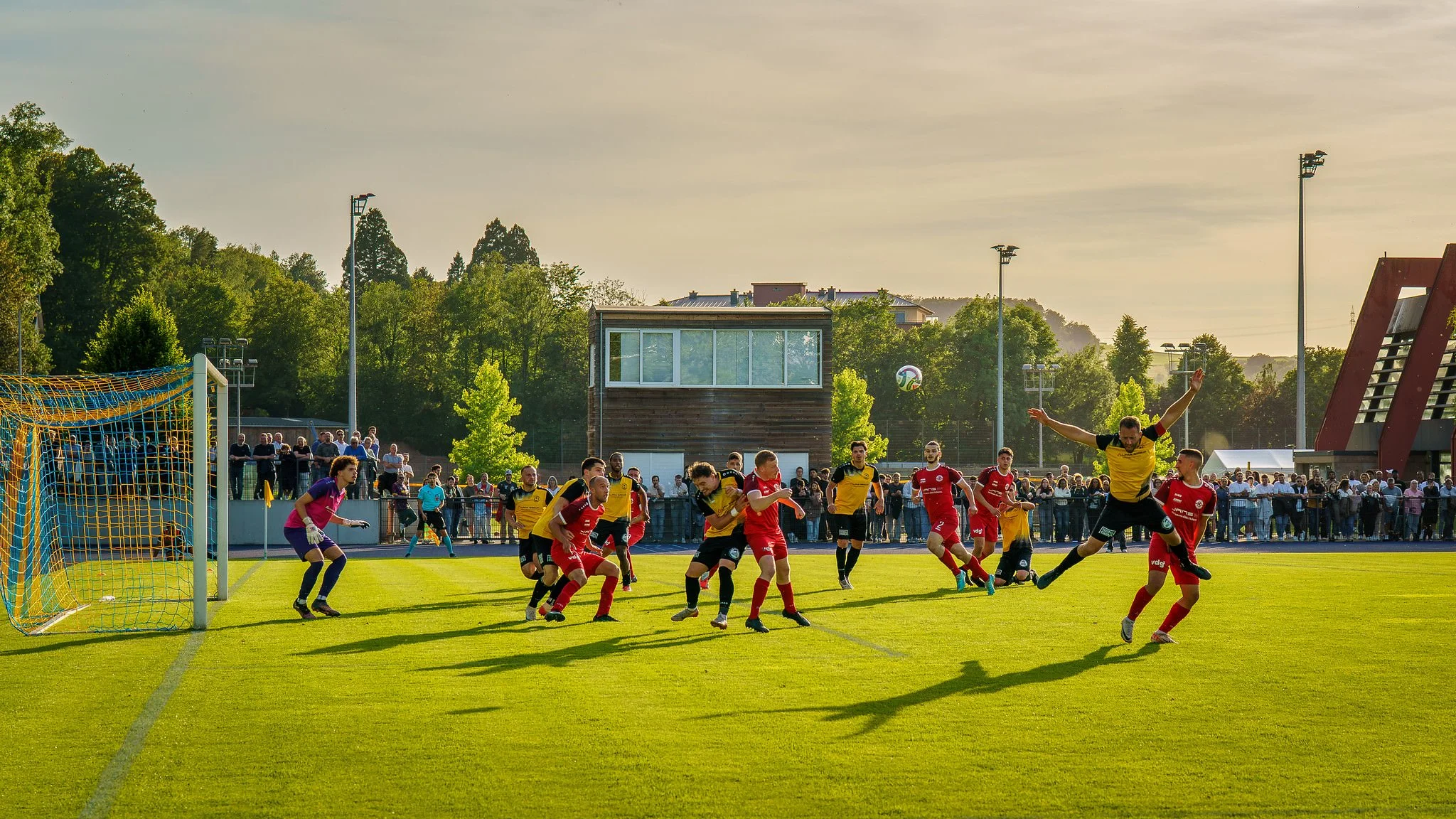 A soccer game in progress on a sunny day, with players in yellow and red uniforms competing near the goal, and spectators watching from the sidelines.