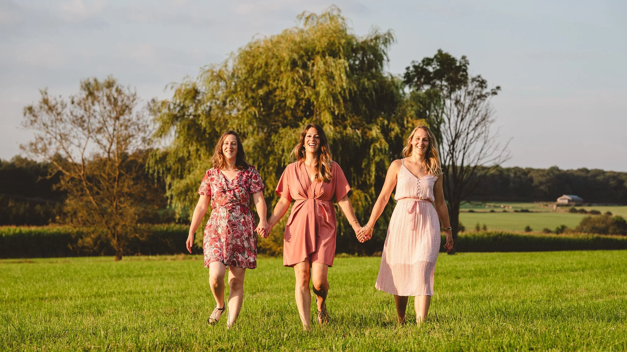 Three sisters wearing dresses holding hands and walking through a grassy field with trees and a barn in the background during daytime. They are enjoying a family photoshoot in Mullerthal, Luxembourg.