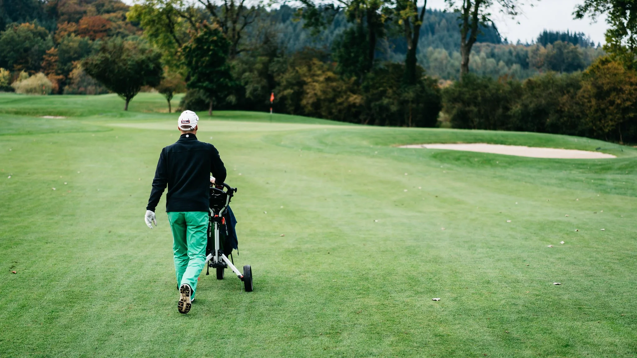 A person walking on a golf course pushing a golf trolley, surrounded by green trees and a sand trap.