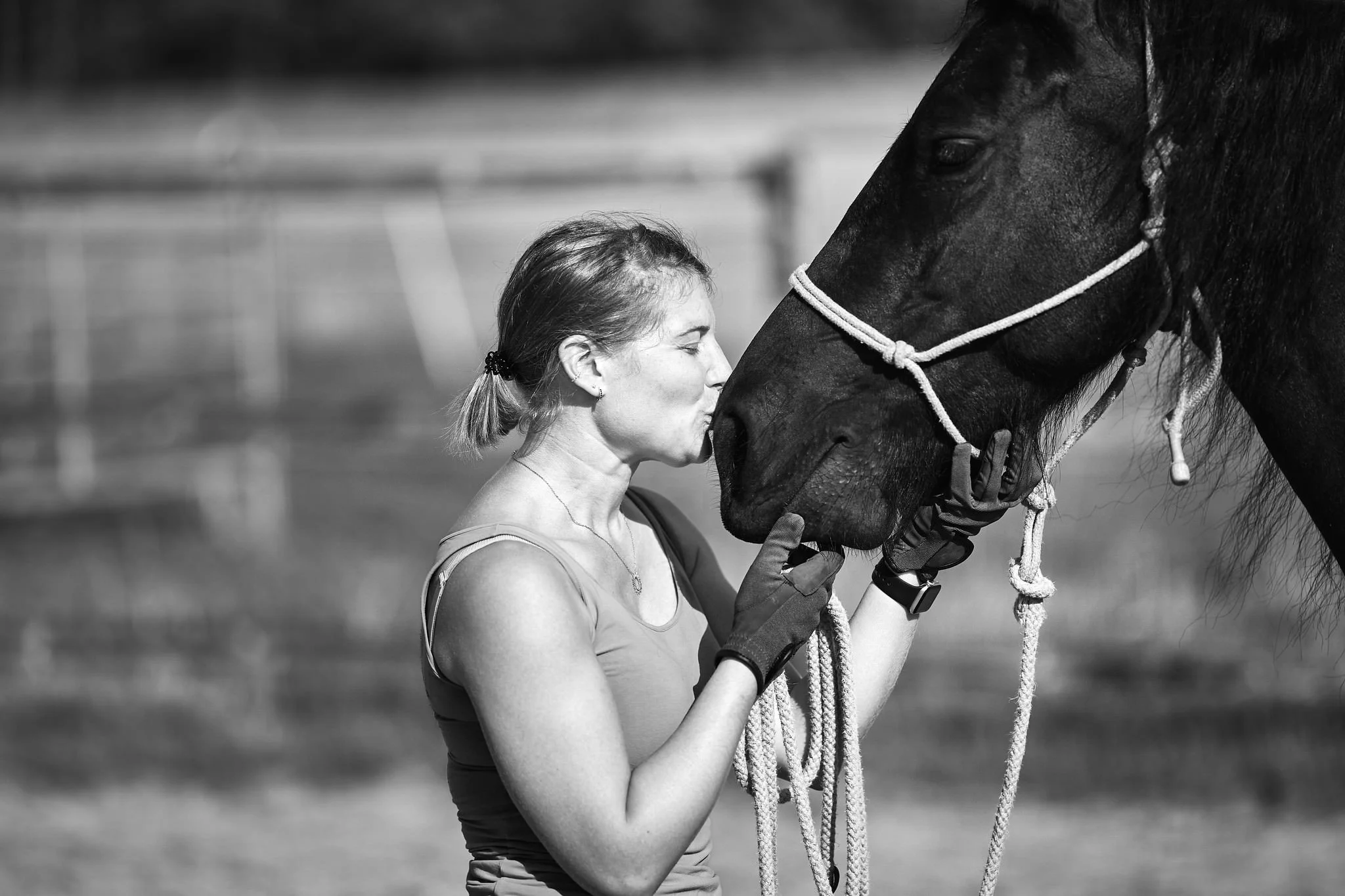 A woman with light hair tied back, wearing a tank top and gloves, kisses a black horse on the face in an outdoor setting.
