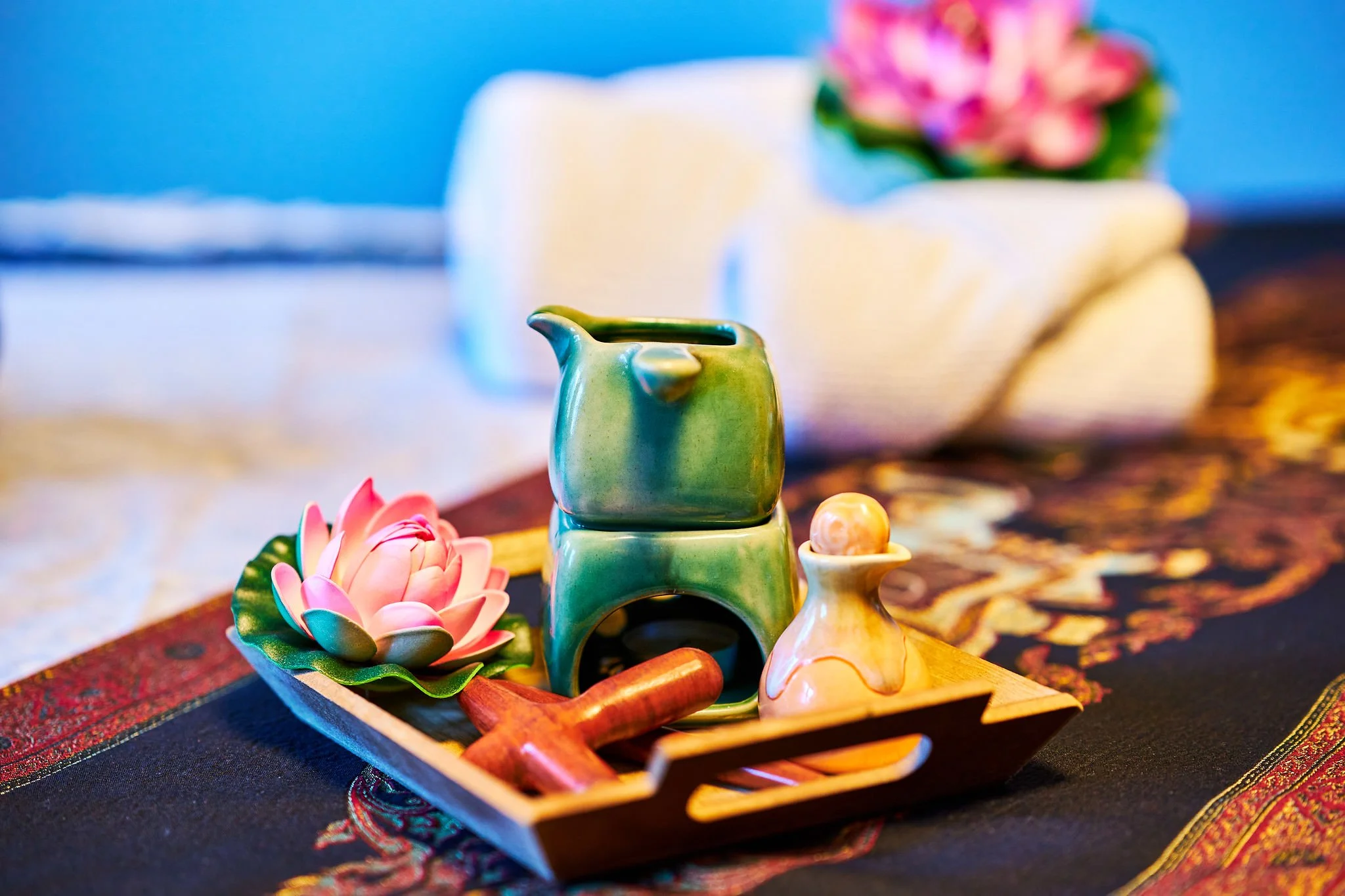 Small green ceramic teapot with a spout, placed on a decorative tray with pink lotus flower and other small ornamental items, against a blurred background with a pink floral arrangement.