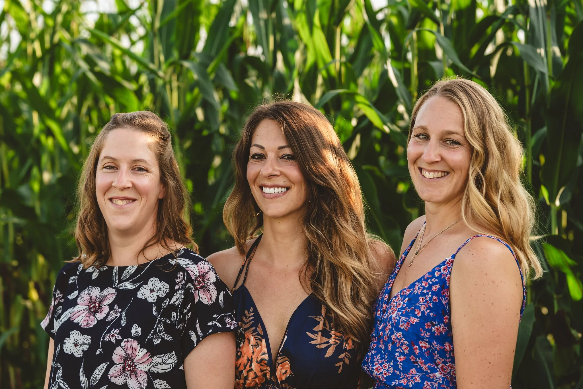 Three sisters smiling outdoors in a green, leafy garden or field. They are enjoying a family photoshoot in Mullerthal, Luxembourg.