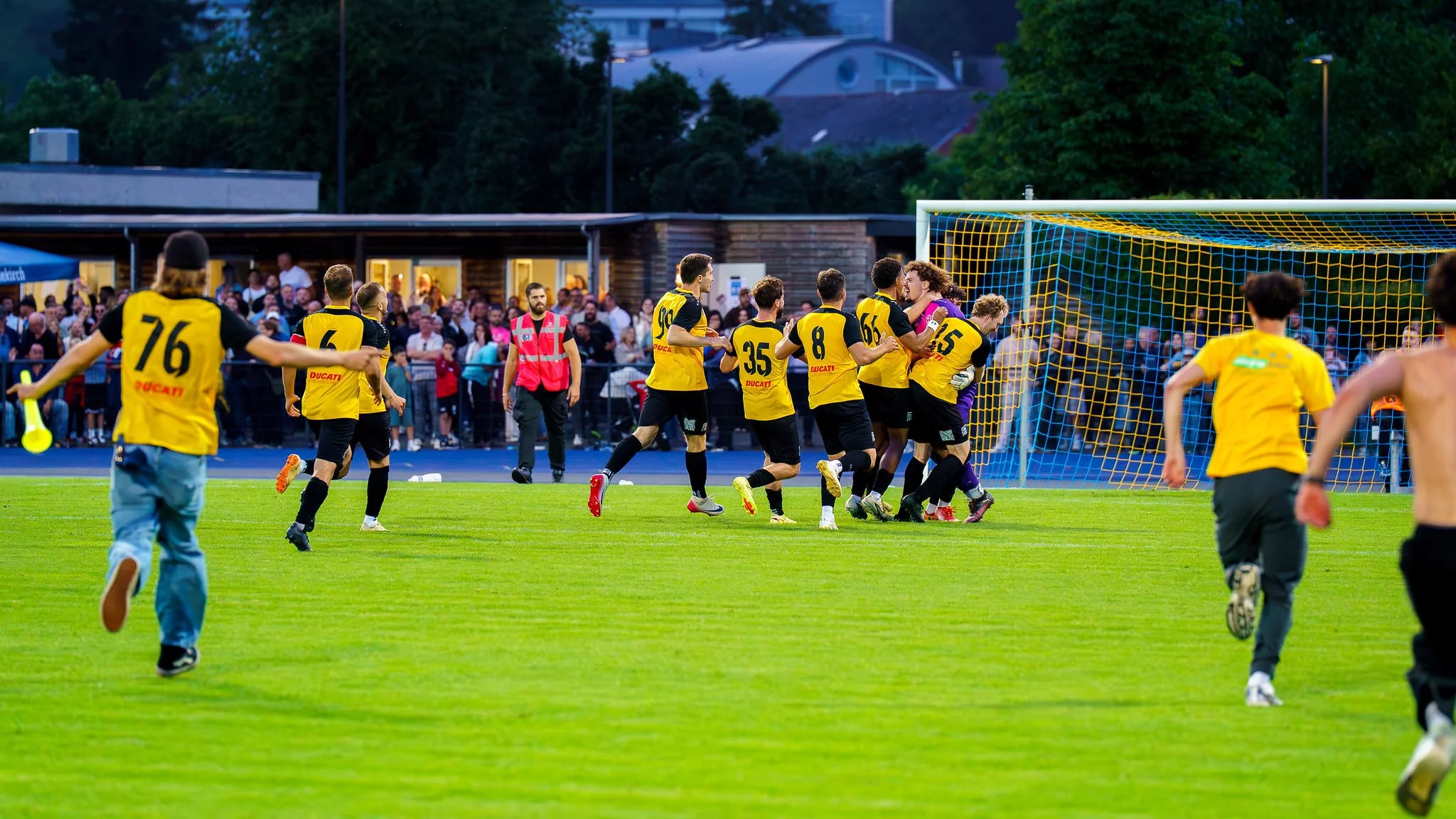 A soccer team in yellow uniforms celebrates near the goalpost after scoring, while spectators watch from the stands.