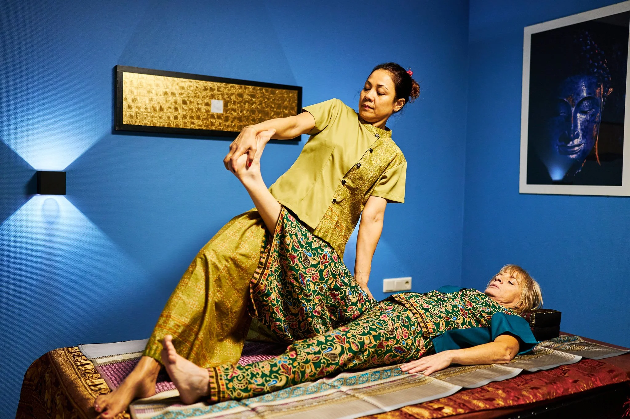 Two women practicing acro yoga in a room with blue walls and Buddha artwork, one woman lying on a mat and the other balancing with her arm around her.