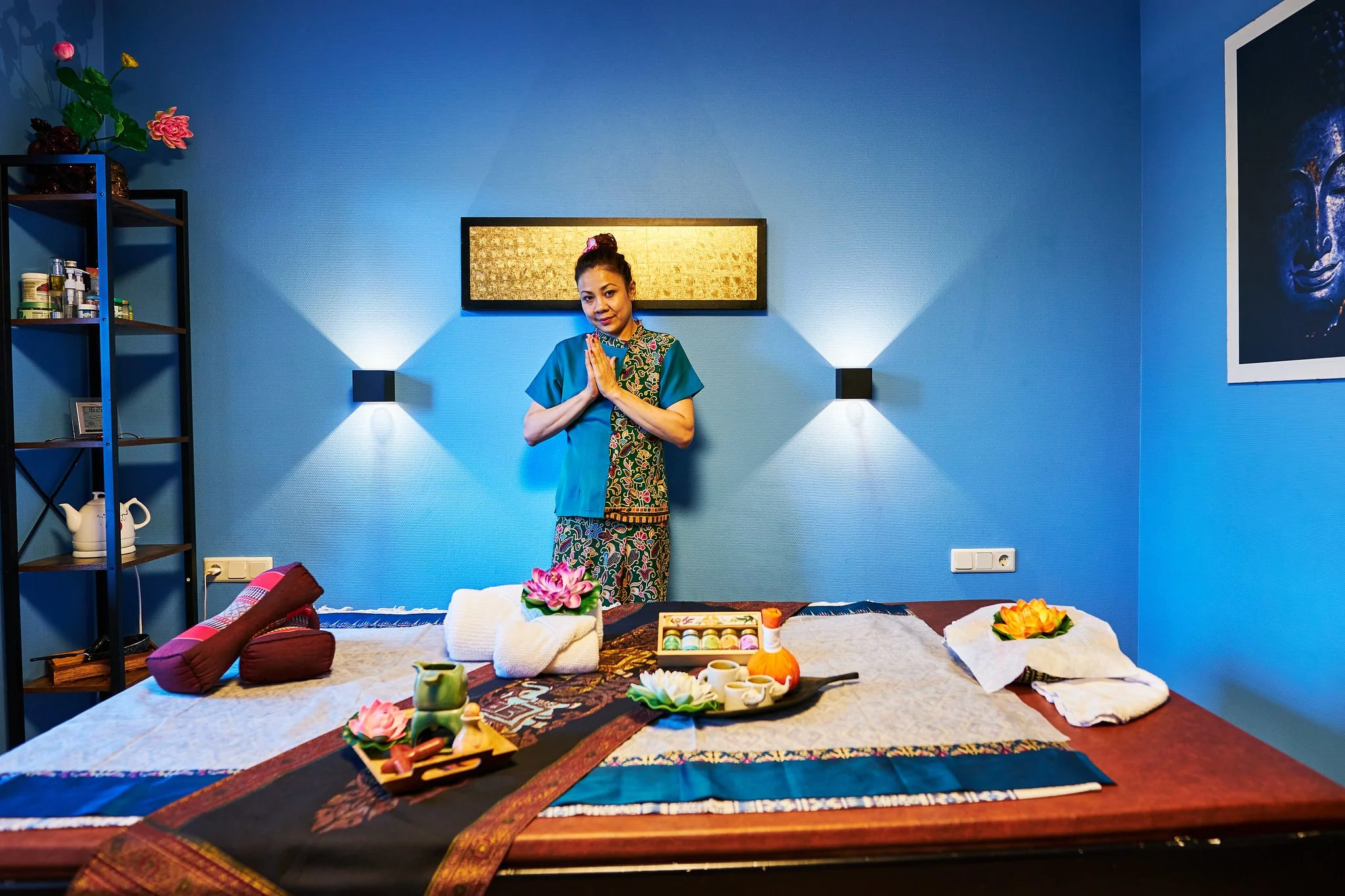 A woman standing in a room with blue walls, hands pressed together in a greeting or prayer gesture. She wears a colorful dress and has her hair in a bun with a pink hair accessory. The room features wall lights, a shelf with various items, a framed a
