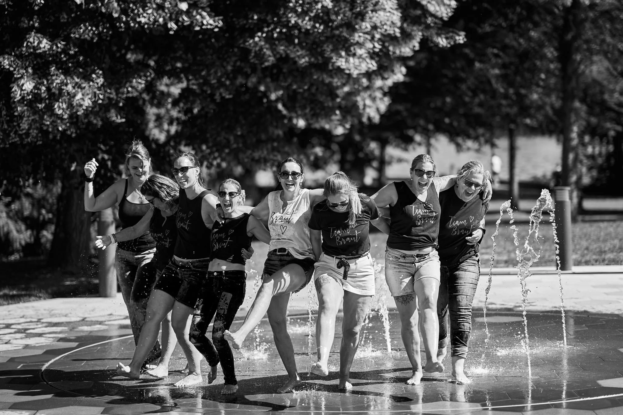 A group of women wearing matching T-shirts with the words 'Team Brad' and heart symbols, laughing and playing in a fountain on a sunny day. They are holding hands and walking through the water splashing around them.
