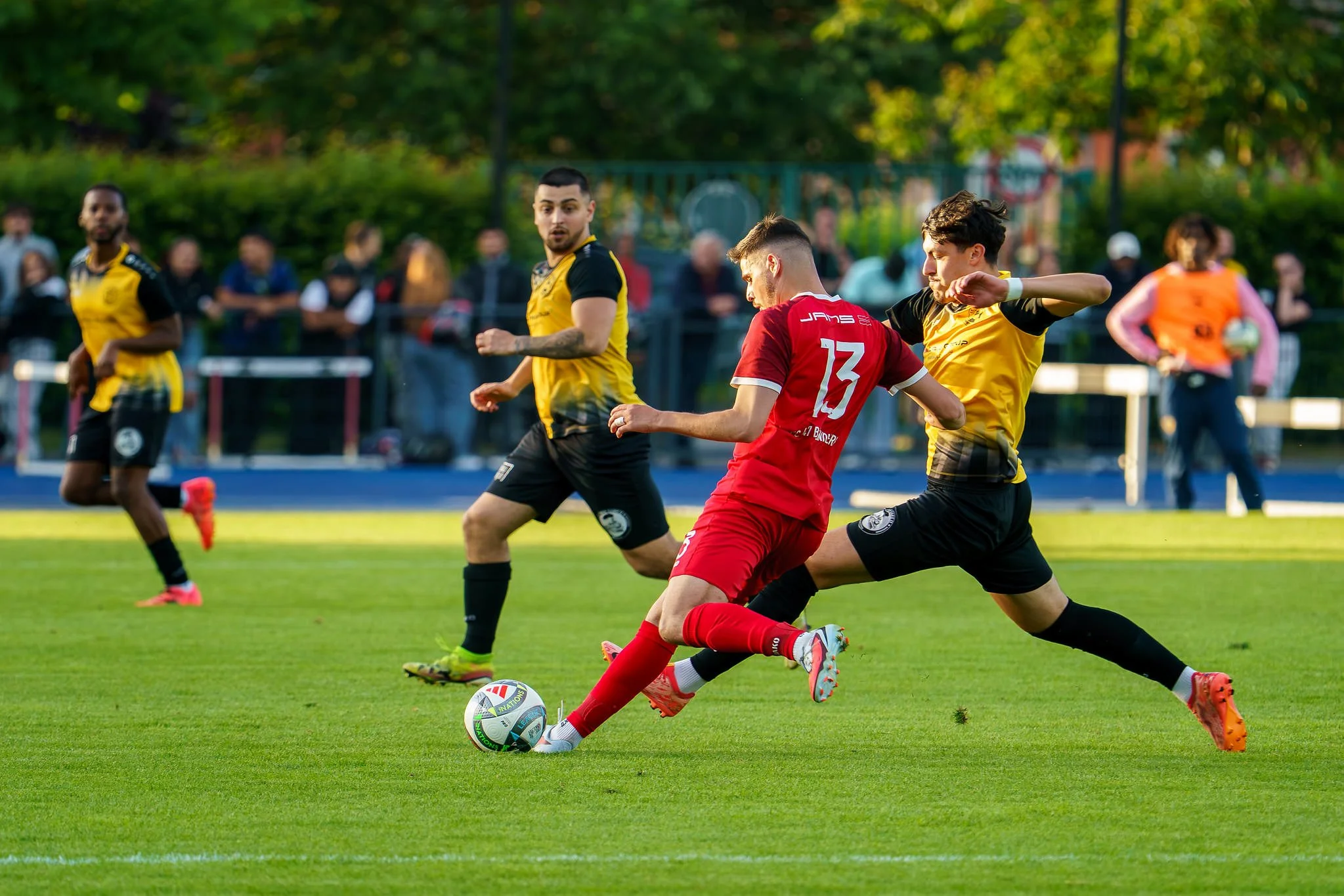 A soccer match in progress with players from two teams, one in red and one in yellow, competing for the ball on a well-maintained grass field with trees in the background. The red team player is kicking the ball while yellow team players are nearby, 
