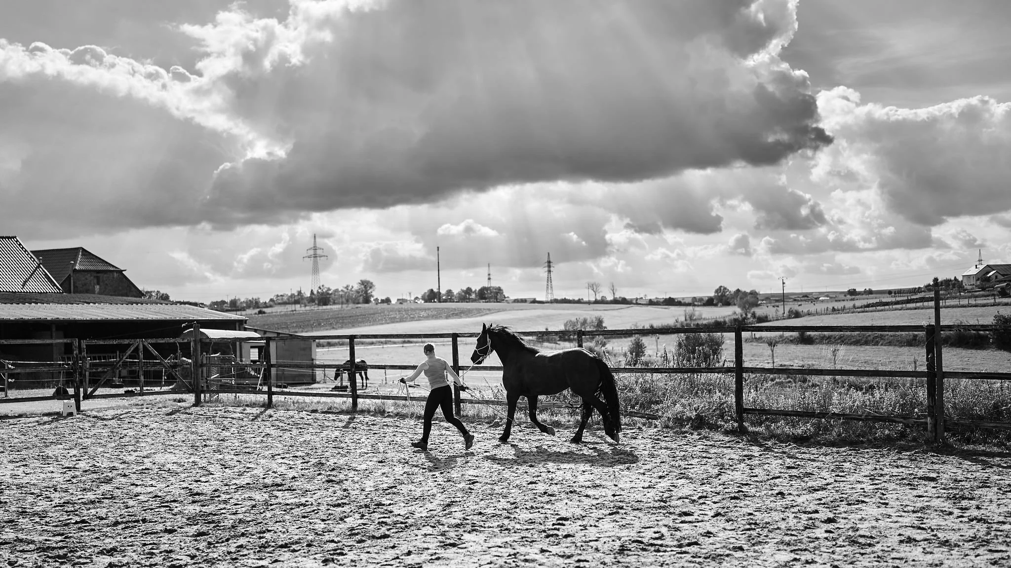 A person leading a horse in a fenced outdoor riding arena with buildings, trees, and power lines in the background under a cloudy sky.