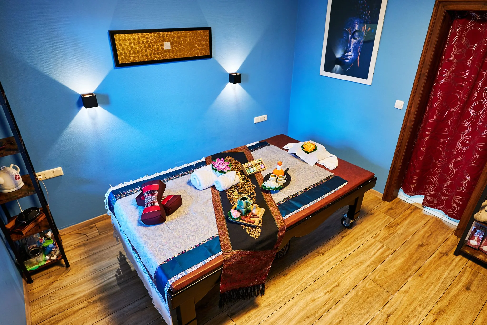 A cozy bedroom with blue walls and wooden floors, featuring a bed with a decorative cover, candles, flowers, and a tray, with wall art and red curtains.