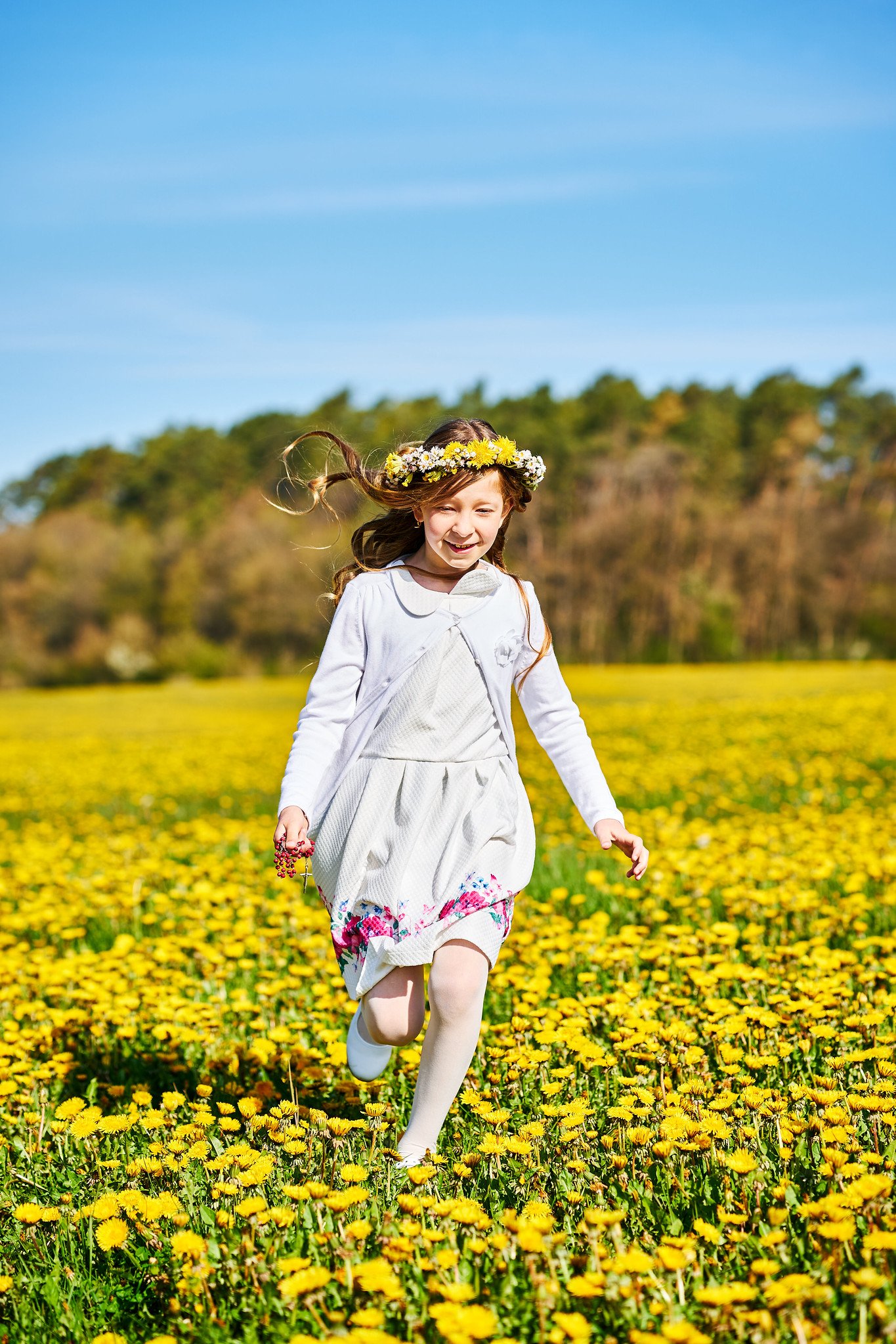 A young girl with long brown hair wearing a white dress and a floral crown runs through a field of yellow flowers under a blue sky.