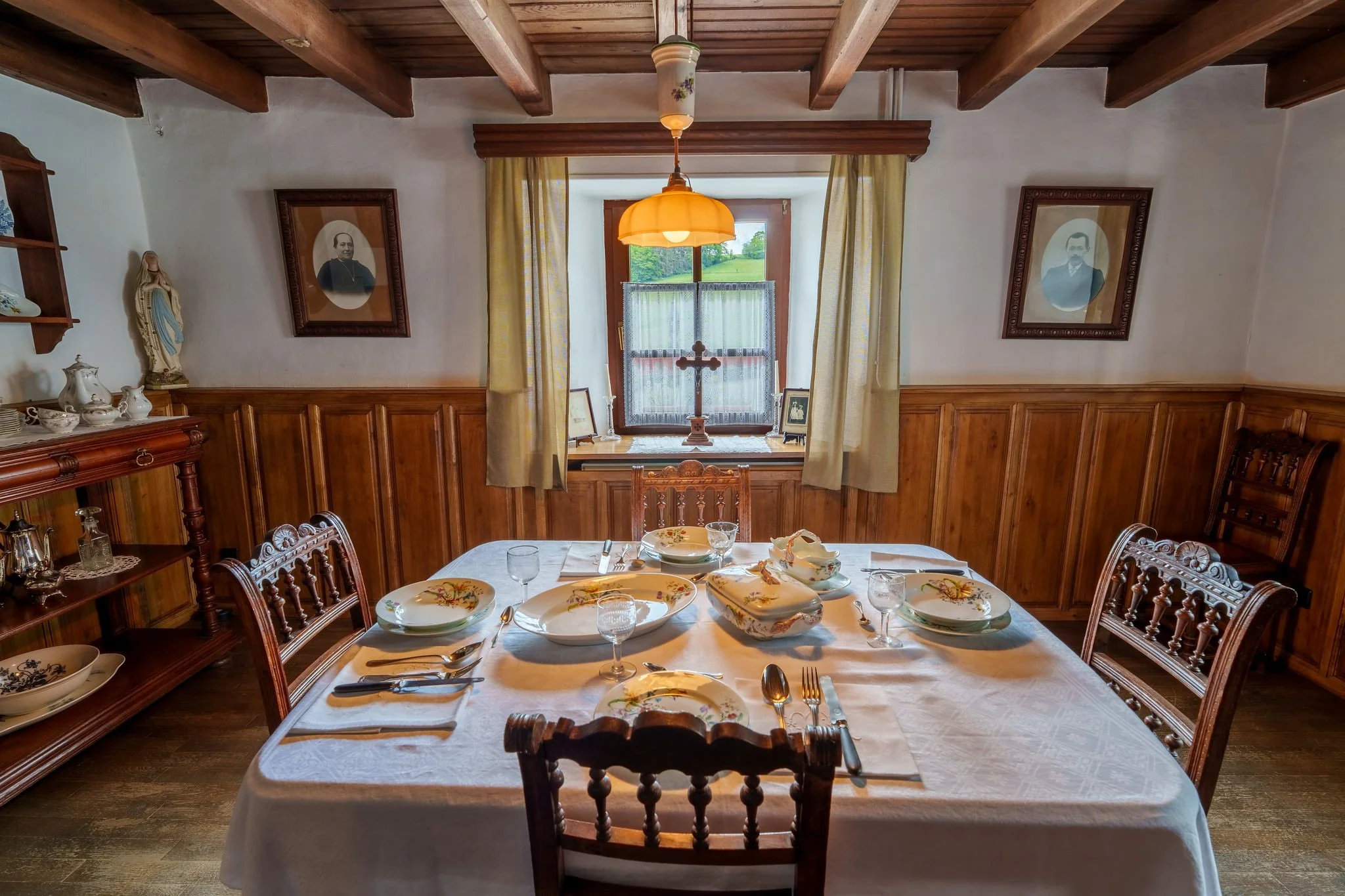 A cozy dining room with a wooden ceiling and wainscoting, a large window with yellow curtains, and a round dining table set with white and floral dishware, silverware, and glasses. The table is covered with a white tablecloth. There are framed black-