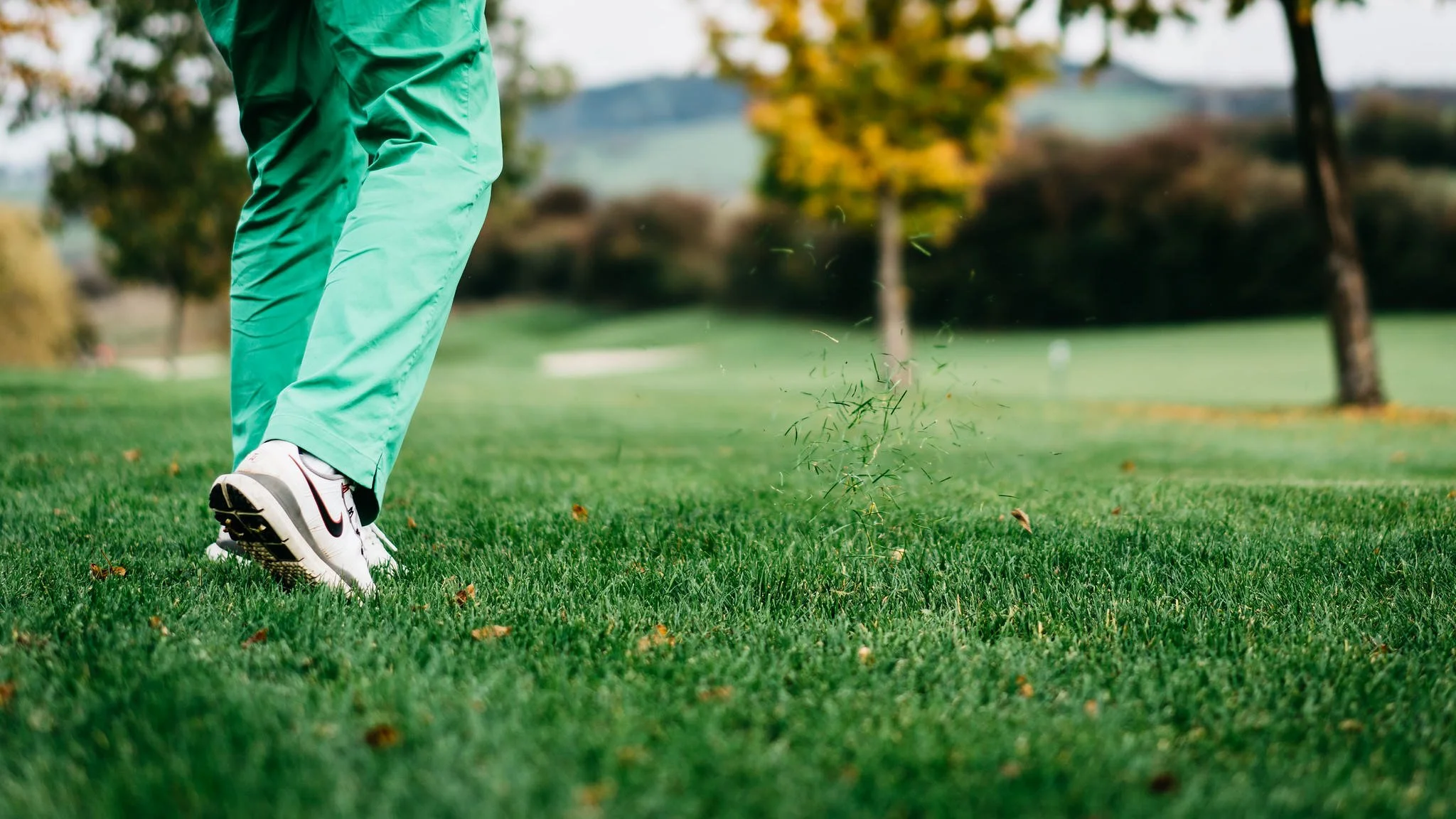 Close-up of a person playing golf on a lush green golf course, focusing on their lower body and golf ball with a background of trees and a slightly cloudy sky.