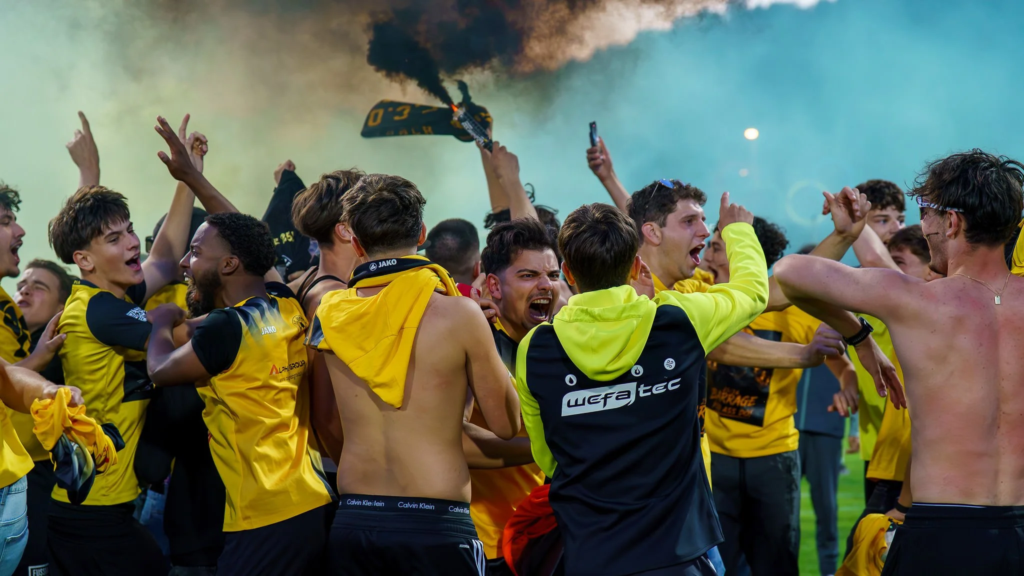 Group of enthusiastic soccer players celebrating after a match, some with arms raised, in a stadium with smoke and fireworks in the background.