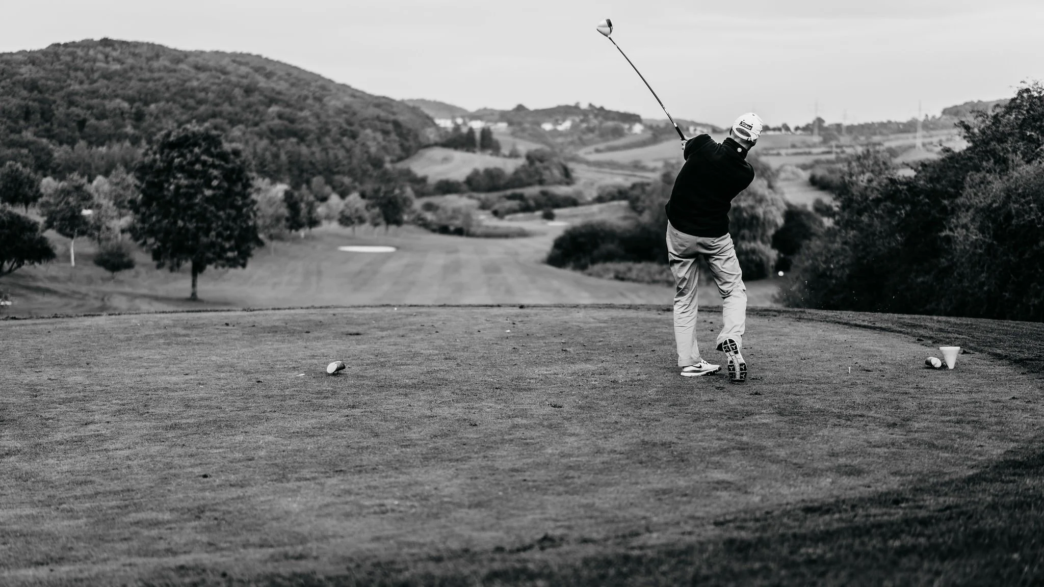 A person playing golf on a course, preparing to hit the ball with a club, with trees and rolling hills in the background.