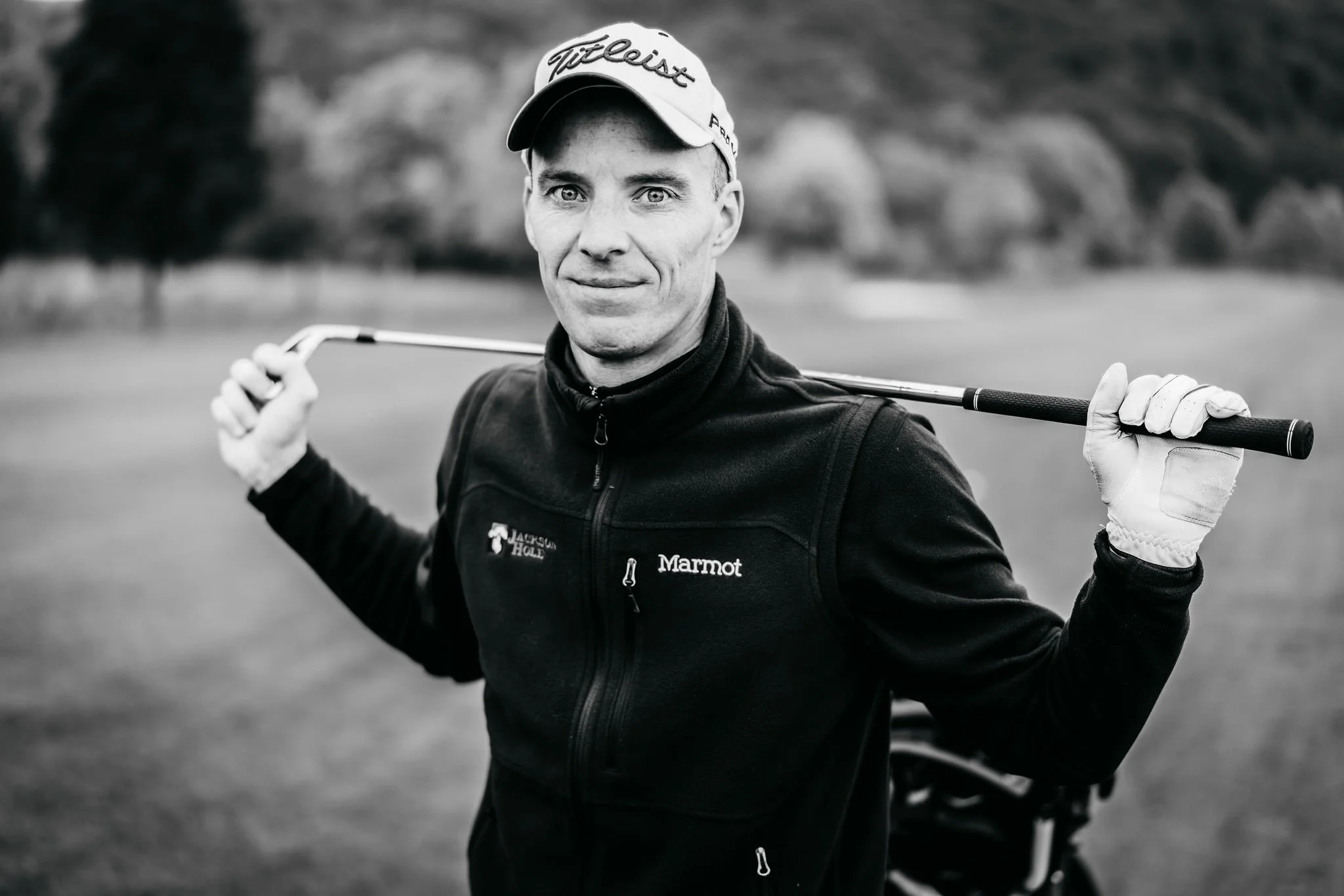 Black and white photo of a man on a golf course holding a golf club behind his neck, wearing a hat and jacket, with trees in the background.