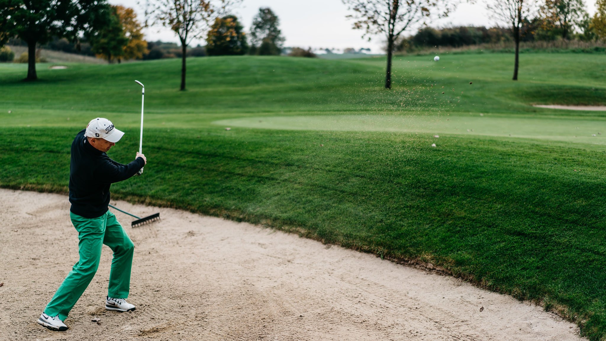A golfer standing in a bunker on a golf course, swinging a club to hit the golf ball towards the green, with trees and a fairway in the background.