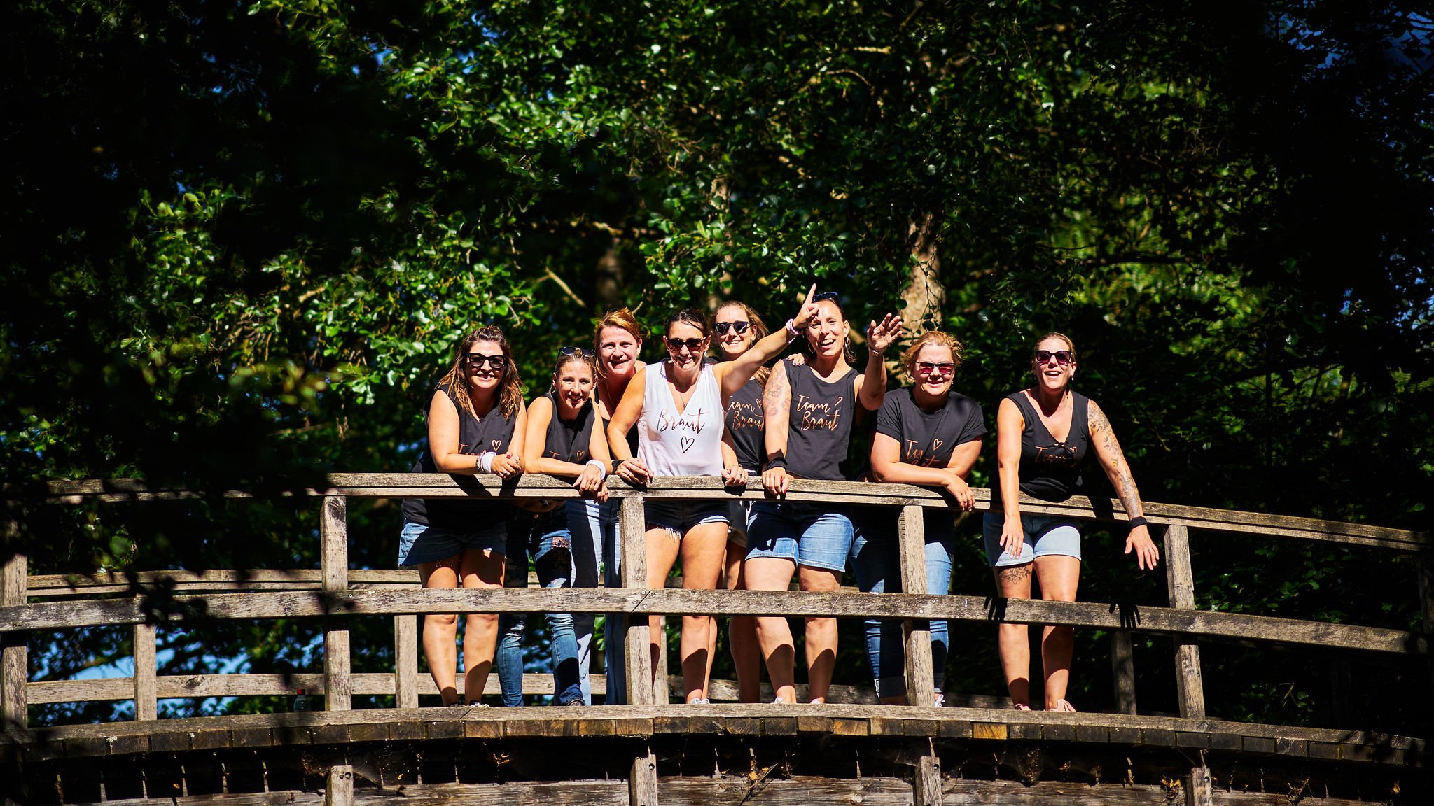 Group of women standing on a wooden bridge outdoors, smiling and posing in front of green trees, wearing casual summer clothing and sunglasses.