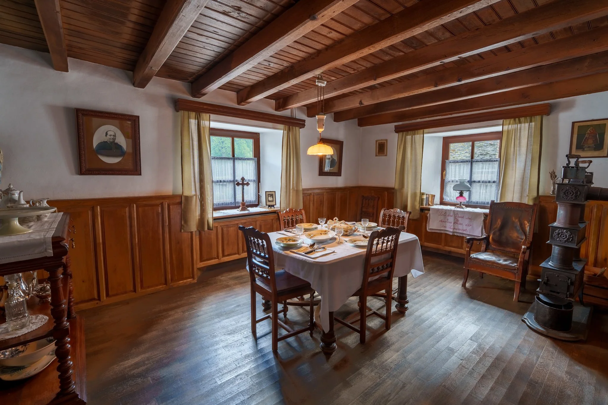 A cozy dining room with wooden ceiling beams and a large window with yellow curtains, set with a table for a meal, surrounded by wooden chairs, and decorated with family portraits and antique items.