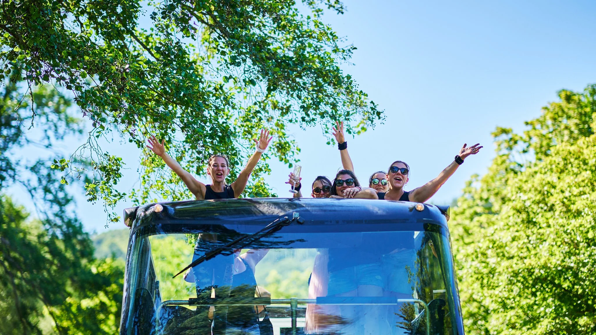 Group of women celebrating and waving at the top of a open vehicle's cabin surrounded by green trees under a clear blue sky.