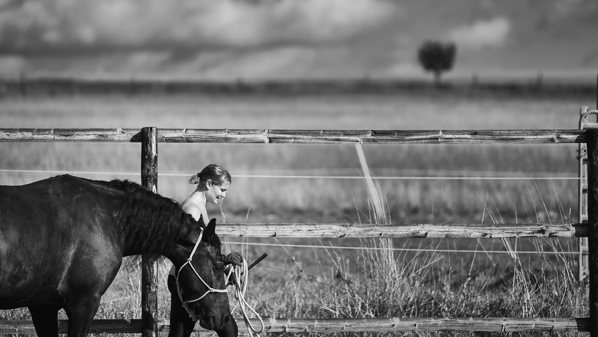 A young woman with a ponytail, wearing a sleeveless top, walking her horse in a rural outdoor setting. The horse is tied to a wooden fence, and the woman is holding a lead rope. The background features open fields, a fence, and a cloudy sky.