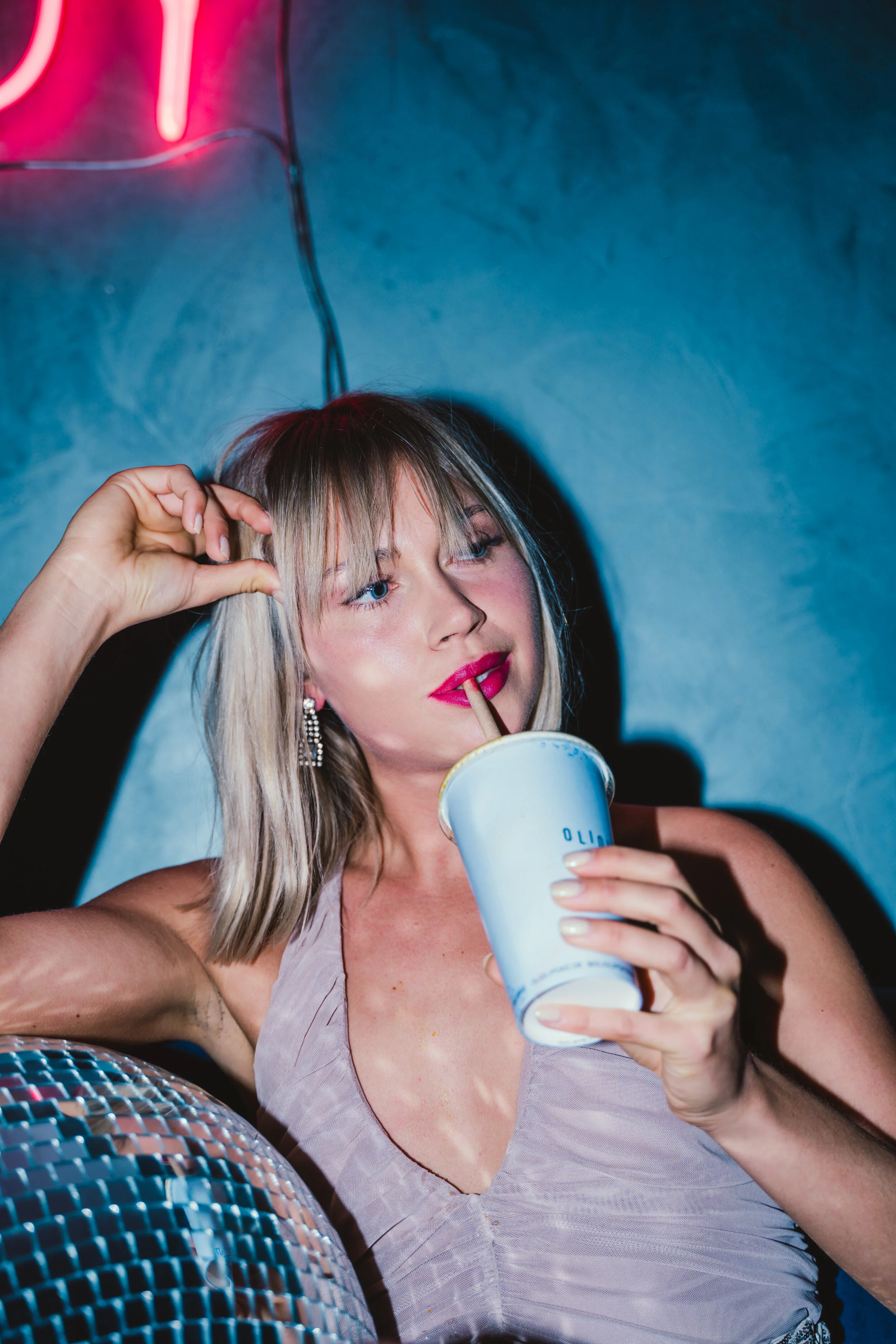 A blonde woman with blue eyes and pink lipstick is holding a drink with a straw, sitting in front of a dark wall and a neon pink sign, with a disco ball in the foreground.