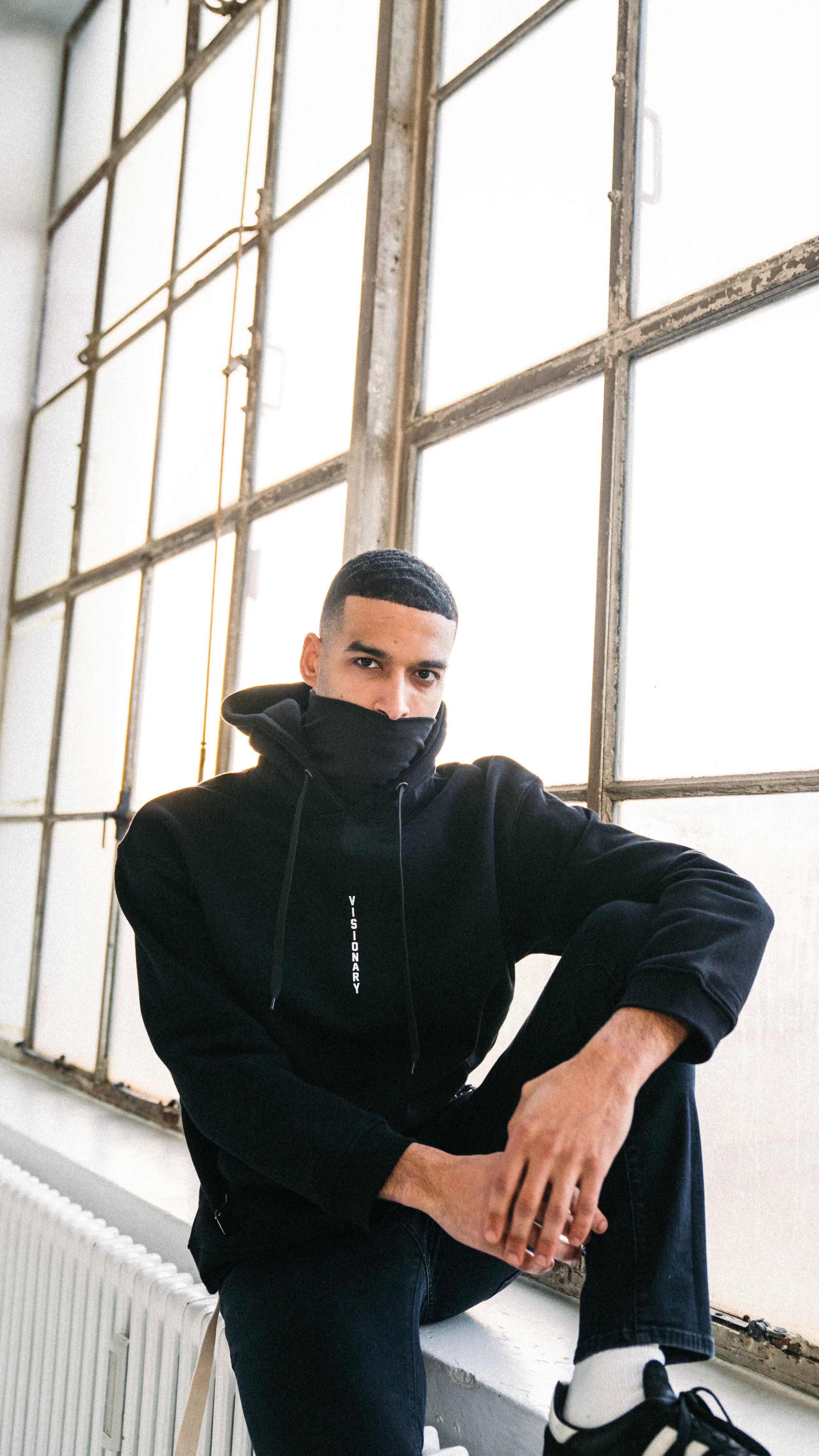A young man sitting on a windowsill in front of large industrial-style windows, wearing a black hoodie with the word 'VISIONARY' on the front, black pants, and black and white sneakers.