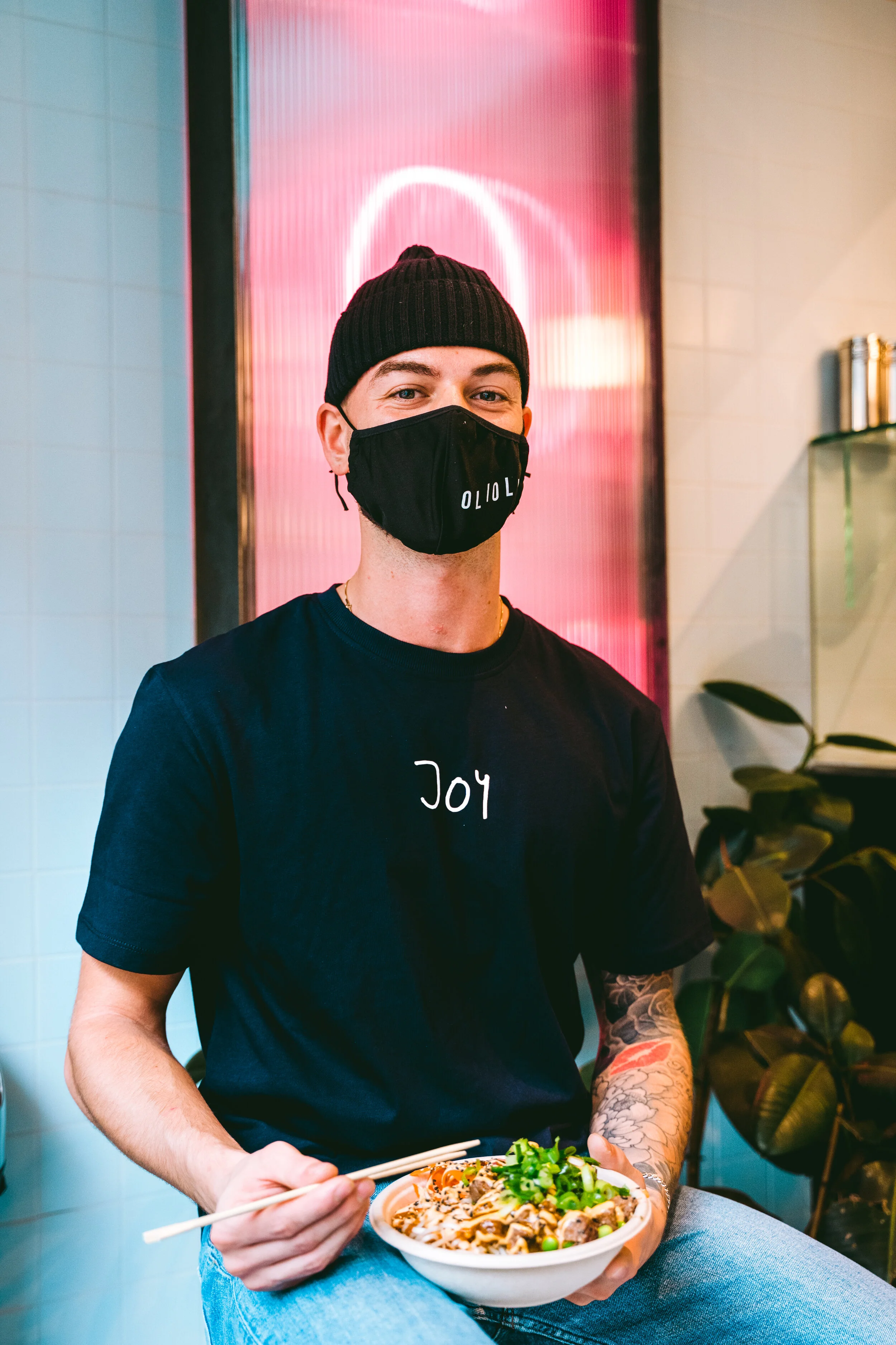 A young man wearing a black beanie and a black face mask sitting in a restaurant, holding a bowl of Asian noodle dish with chopsticks, with a tattoo on his left arm and casually dressed in a black t-shirt that says 'Joy'.