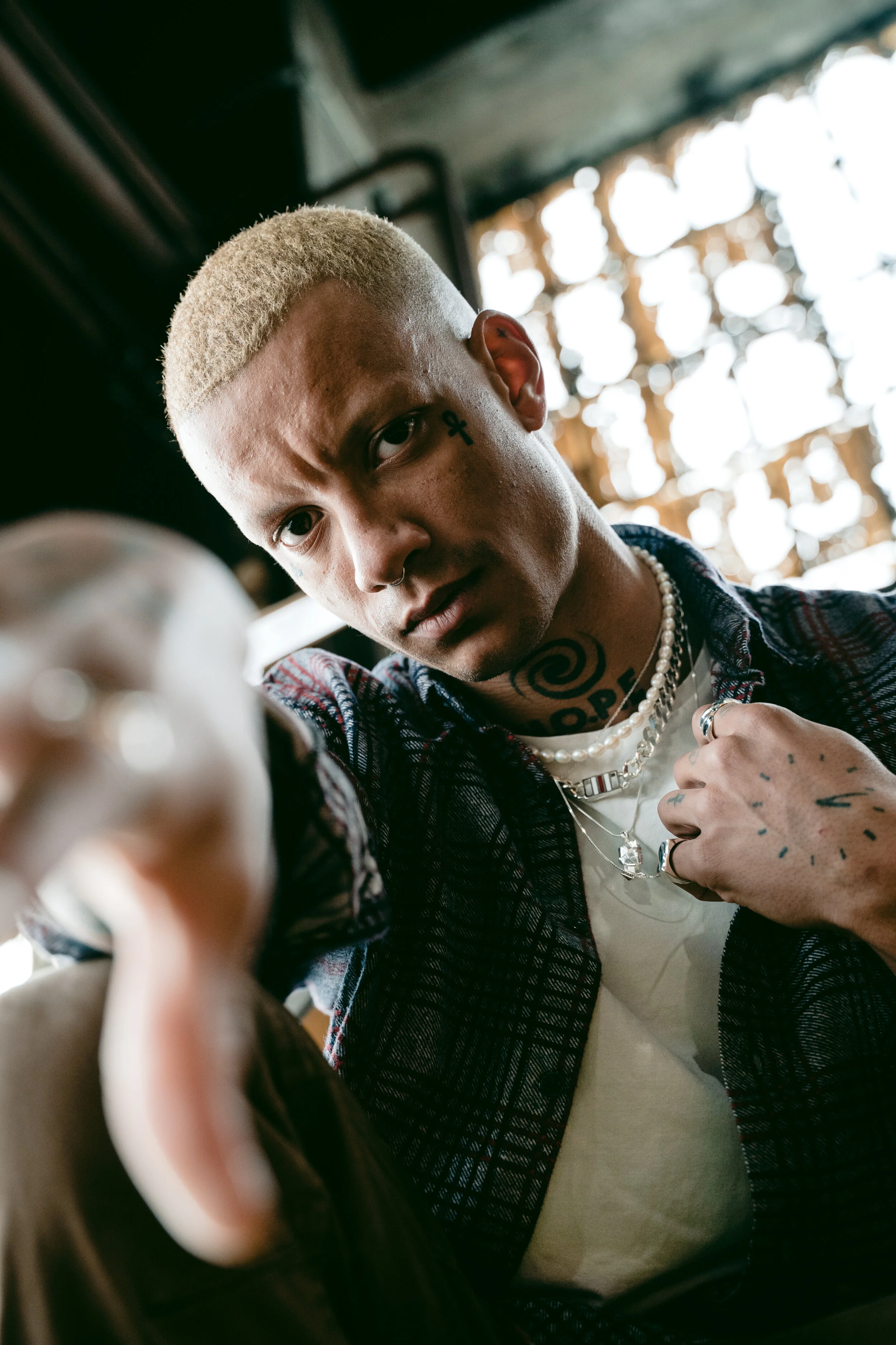 Young man with bleached blond hair, tattoos, and piercings, looking directly at the camera inside a dimly lit space with natural light coming through a window.