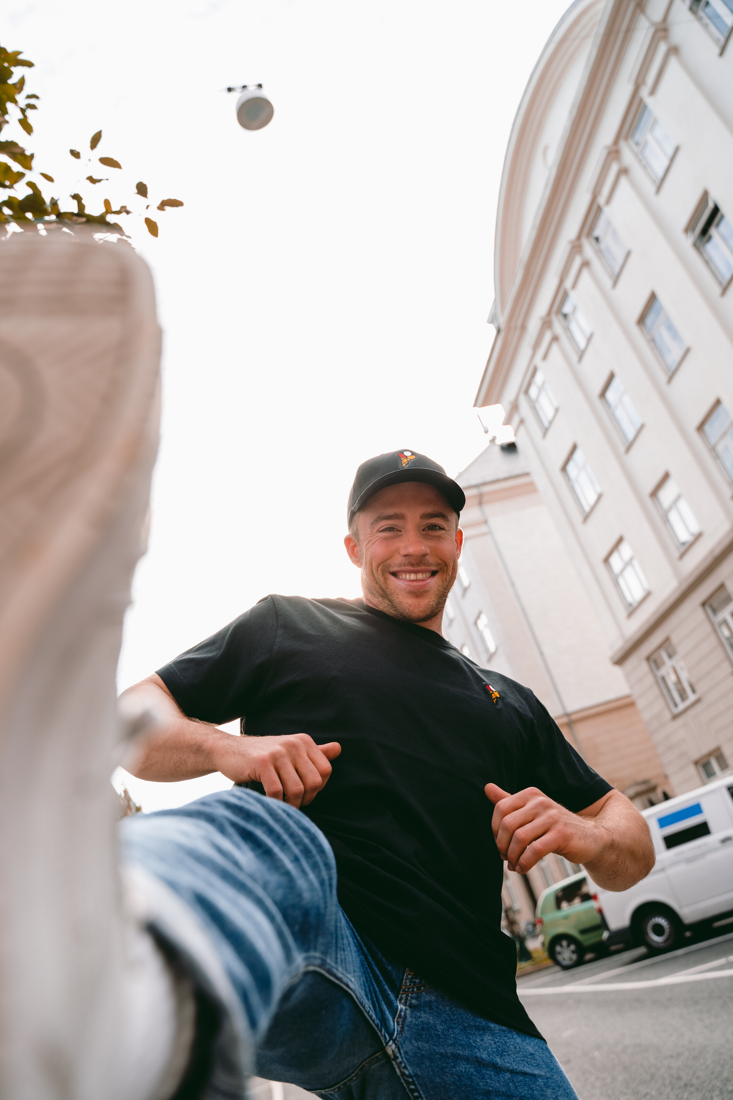 A young man with a broad smile, wearing a black t-shirt and a black cap, is seen from a low-angle view on a city street with tall buildings in the background; his foot is in the foreground, suggesting he is kicking or stepping forward.