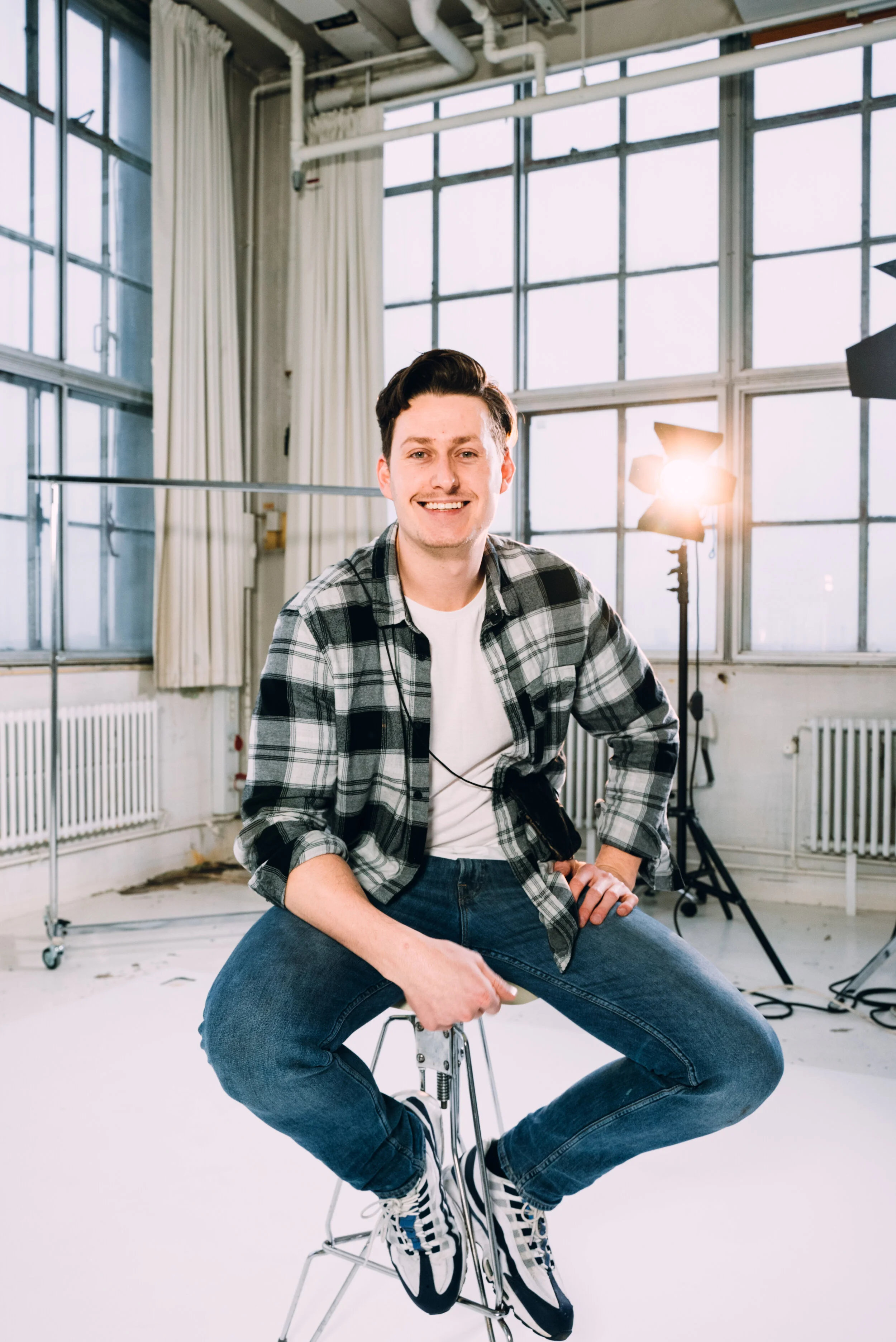 A young man with dark hair, wearing a plaid shirt and jeans, sitting on a stool in a photography studio with large windows, studio lights, and equipment.