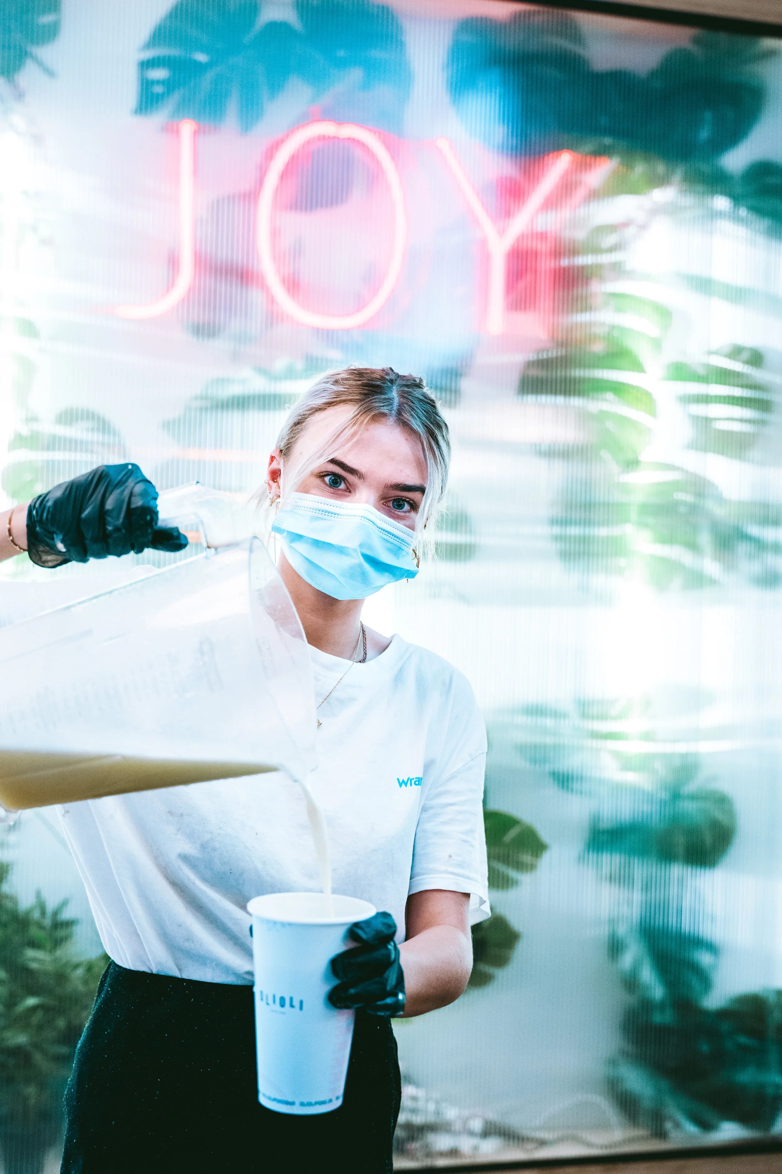 A woman wearing a face mask and black gloves pours a beverage into a cup in front of a green and pink tropical-themed neon sign that reads 'JOY'.