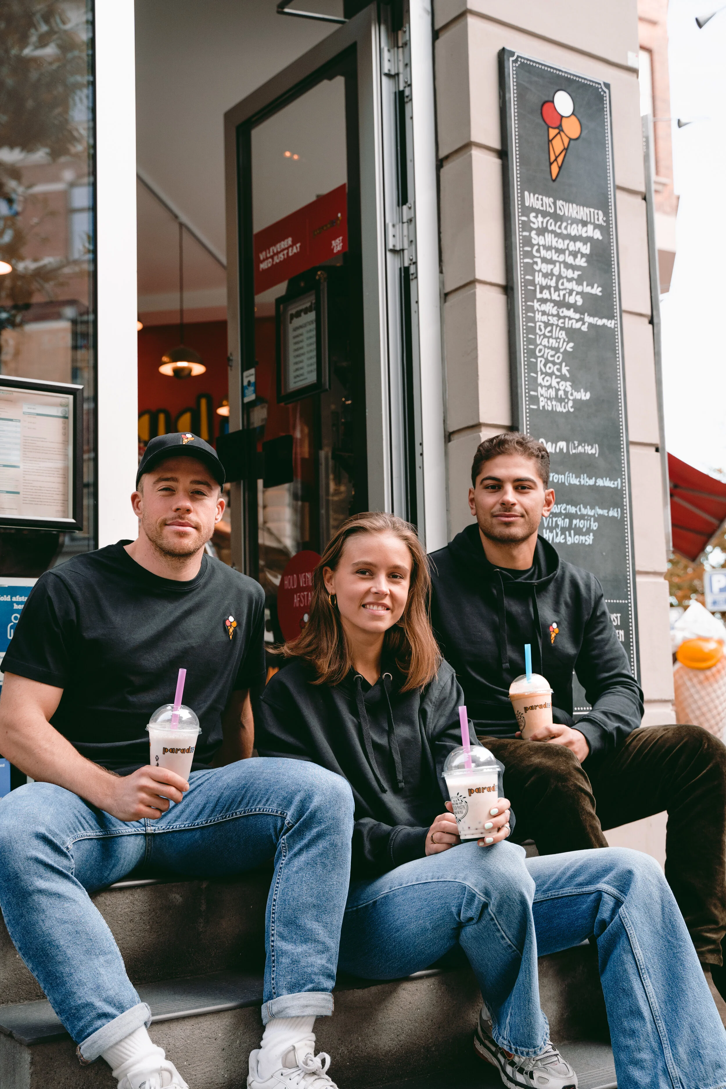 Three young people sitting on a step outside an ice cream shop, holding cups of ice cream with colorful straws, smiling and looking at the camera.