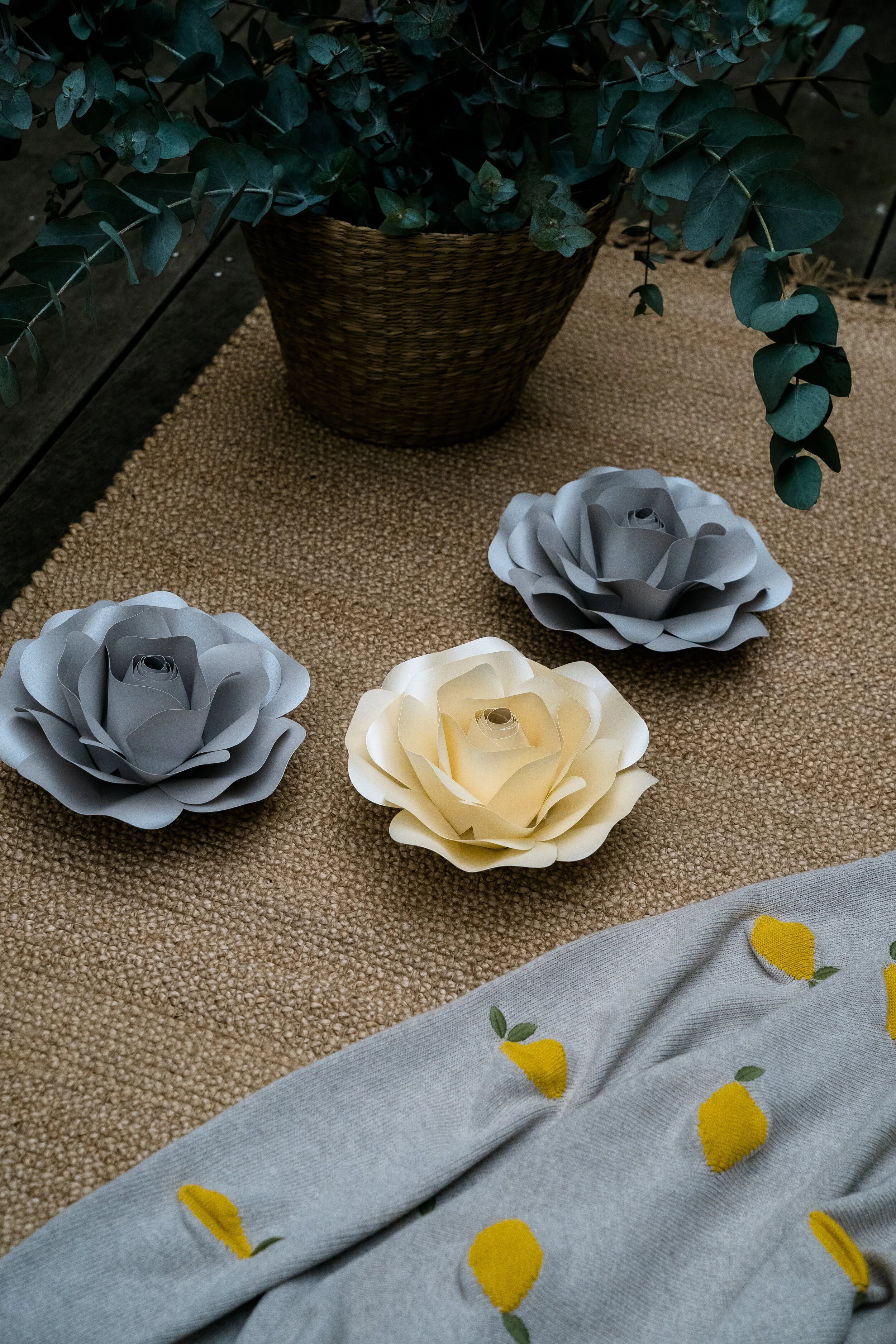 Three large paper roses, two gray and one cream, on a textured beige surface under a potted eucalyptus plant with dark green leaves.