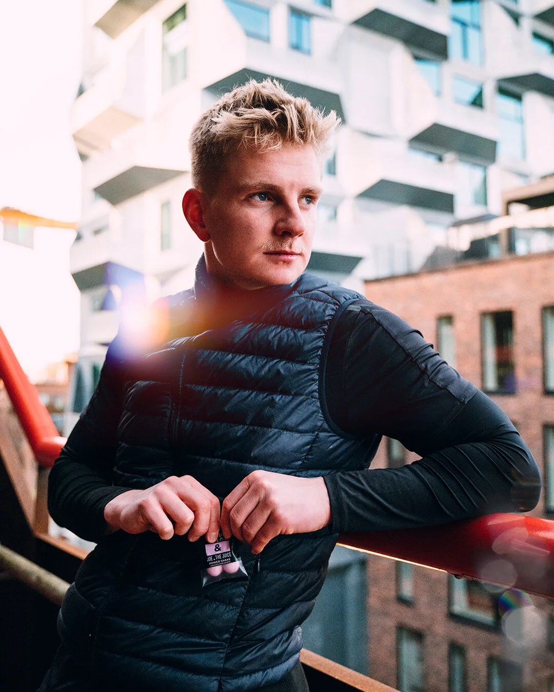 A young man with blonde hair and blue eyes stands on a balcony, holding a packaged juice box in front of a modern building with multiple balconies.