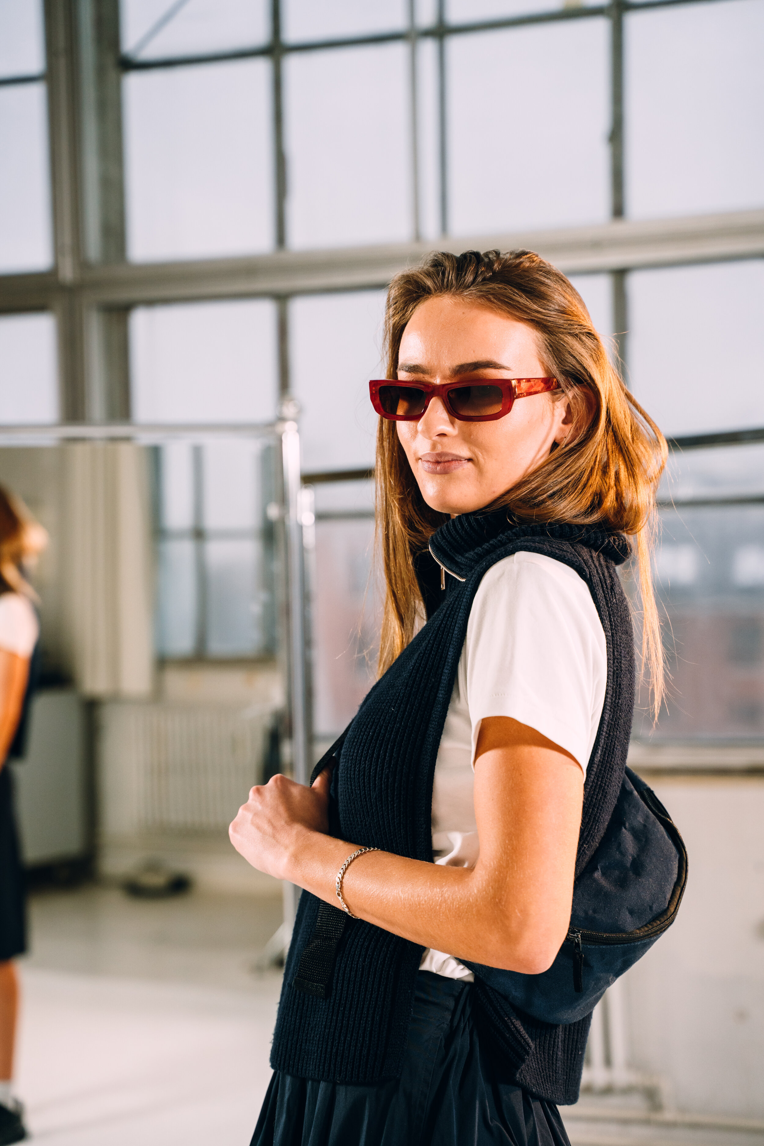 A woman with red hair and sunglasses at an indoor location with large windows, wearing a white T-shirt and a black vest, holding a black backpack over her shoulder.