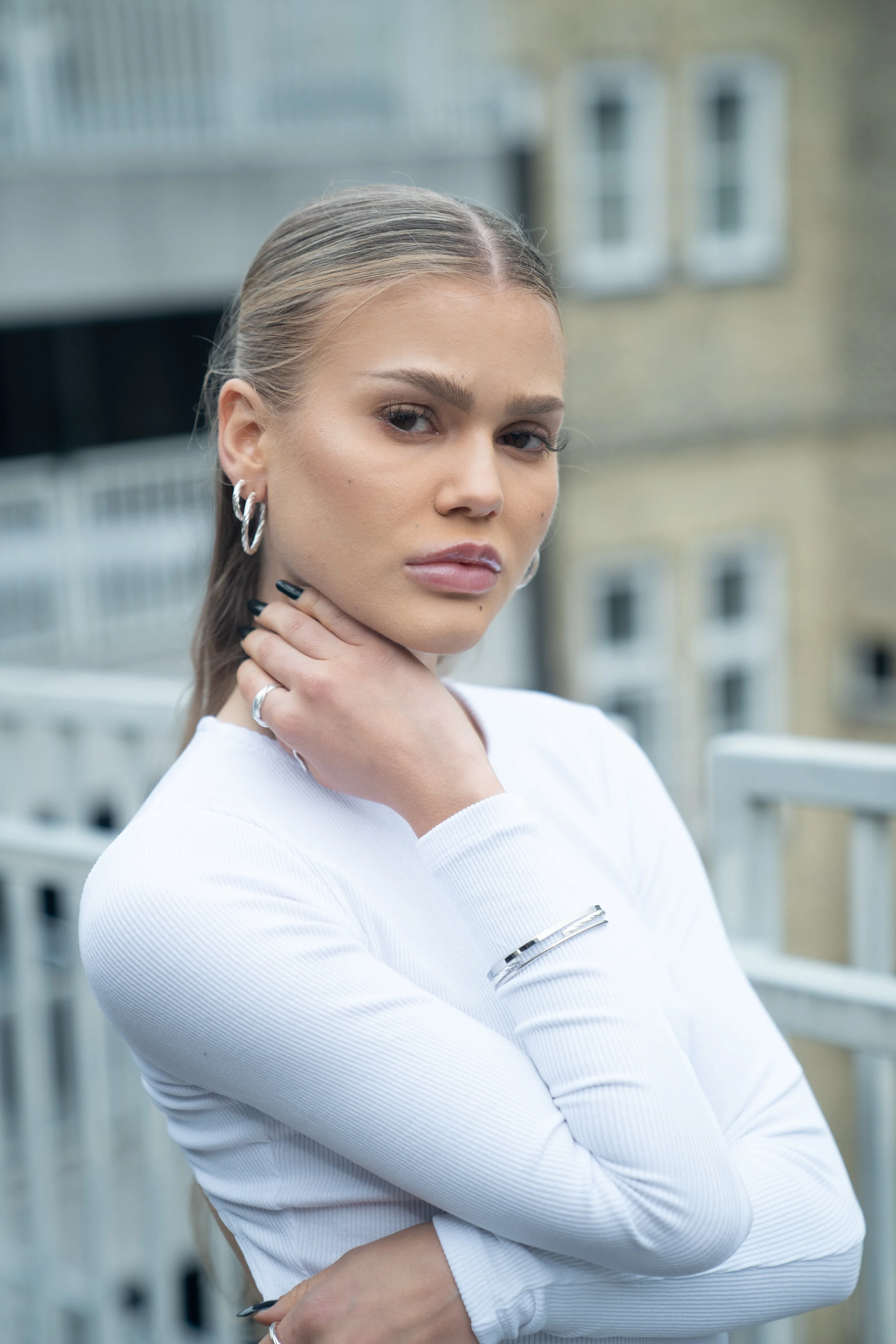 A young woman with blonde hair in a ponytail, wearing a white long-sleeve top and silver jewelry, stands outdoors with an urban building background.