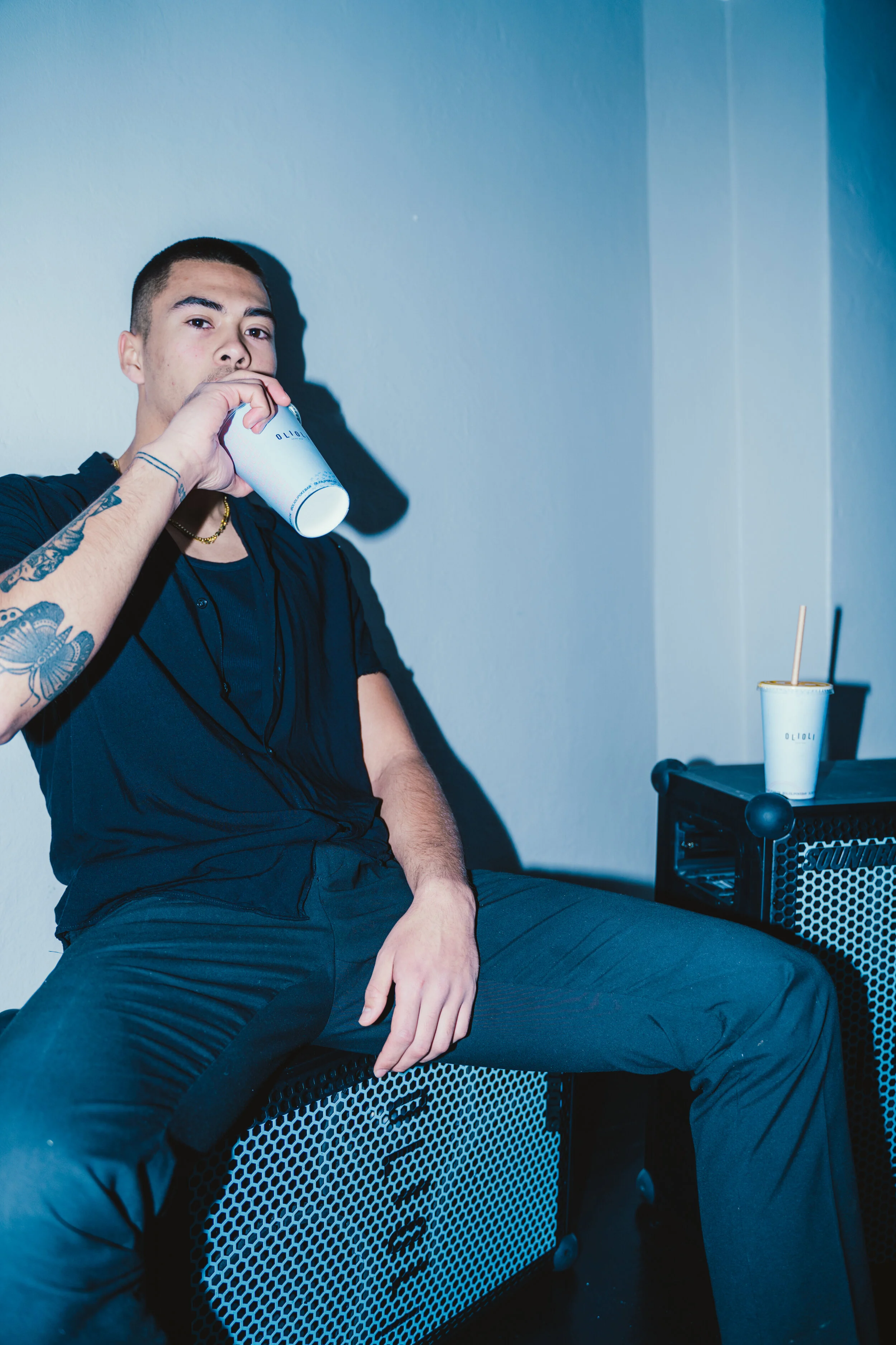 A young man with tattoos on his arm sits on a speaker, drinking from a white cup, with another similar cup on a nearby table.