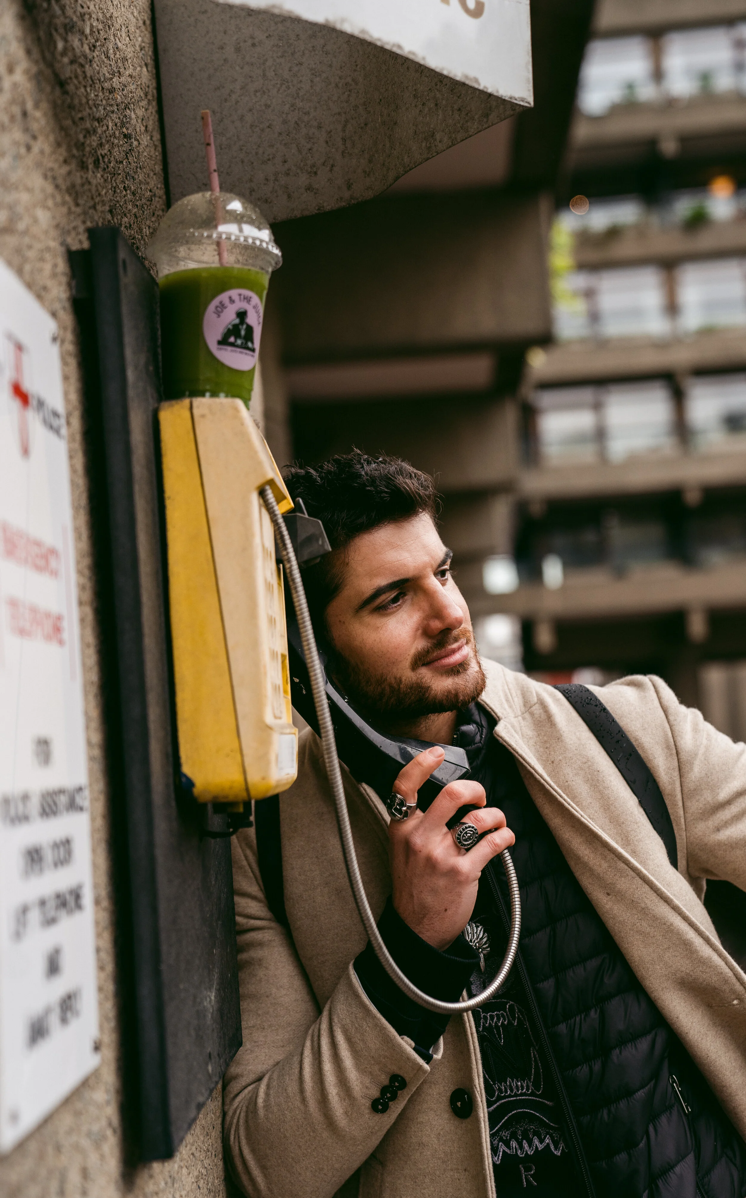 A man with dark hair and beard talking on a public payphone attached to a wall, with a green smoothie in a clear plastic cup placed above the payphone.