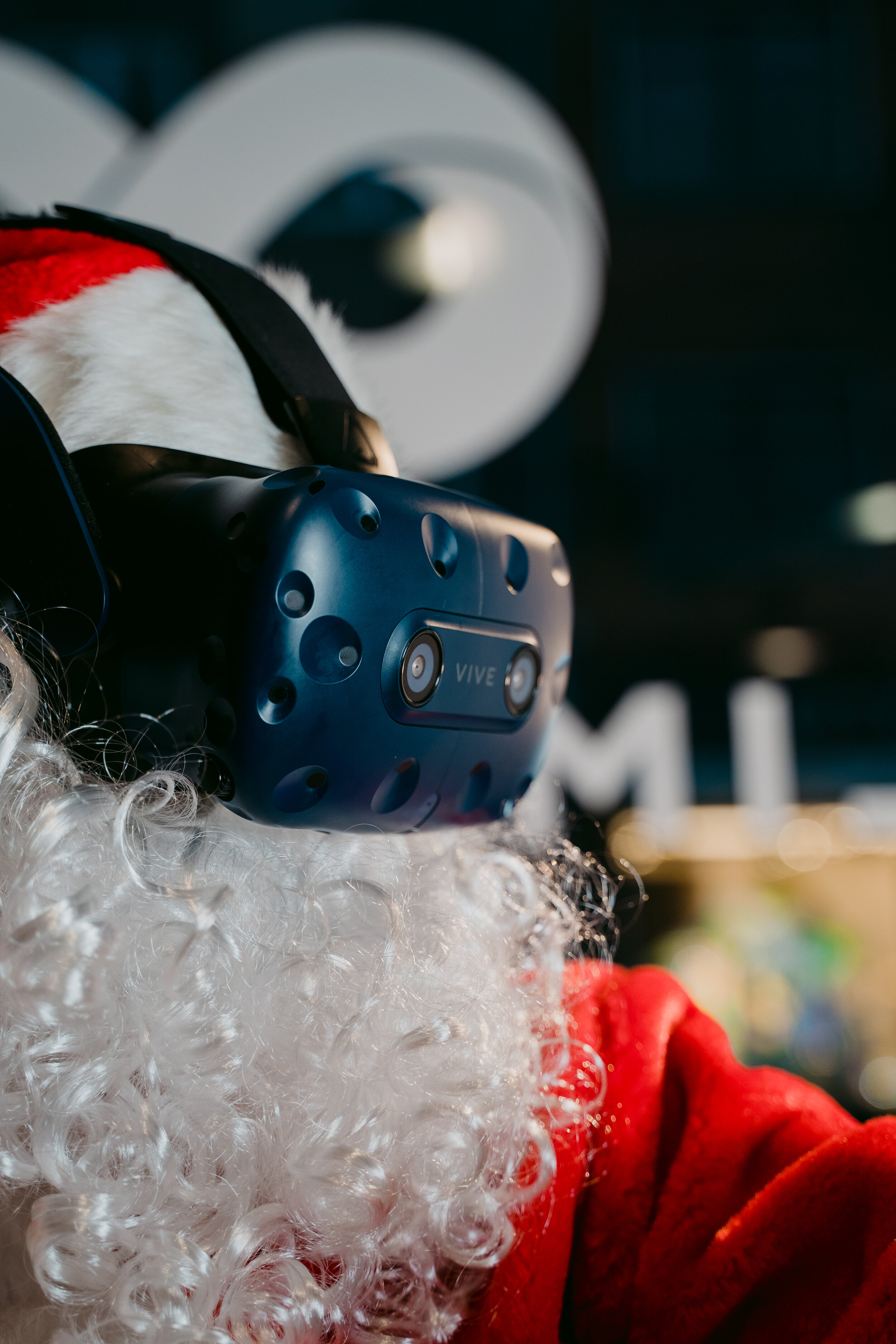 Person dressed as Santa Claus wearing a virtual reality headset, with Christmas hat and curly white beard, in an indoor setting.