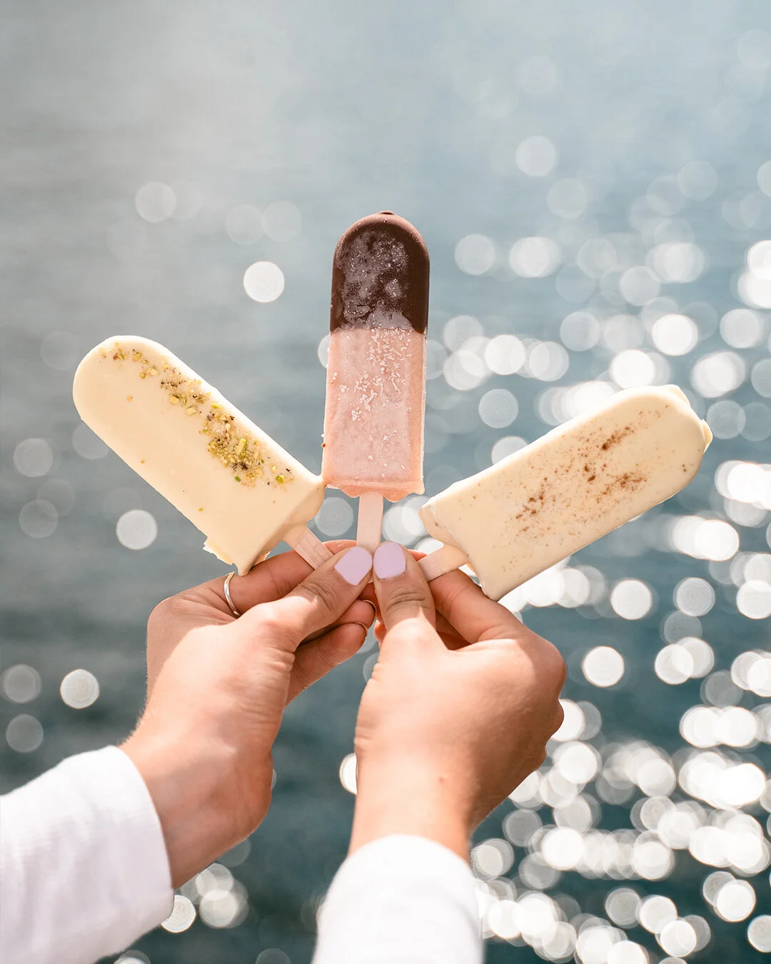Three ice cream bars held in front of a sparkling water background, with a hand showing pink nails.