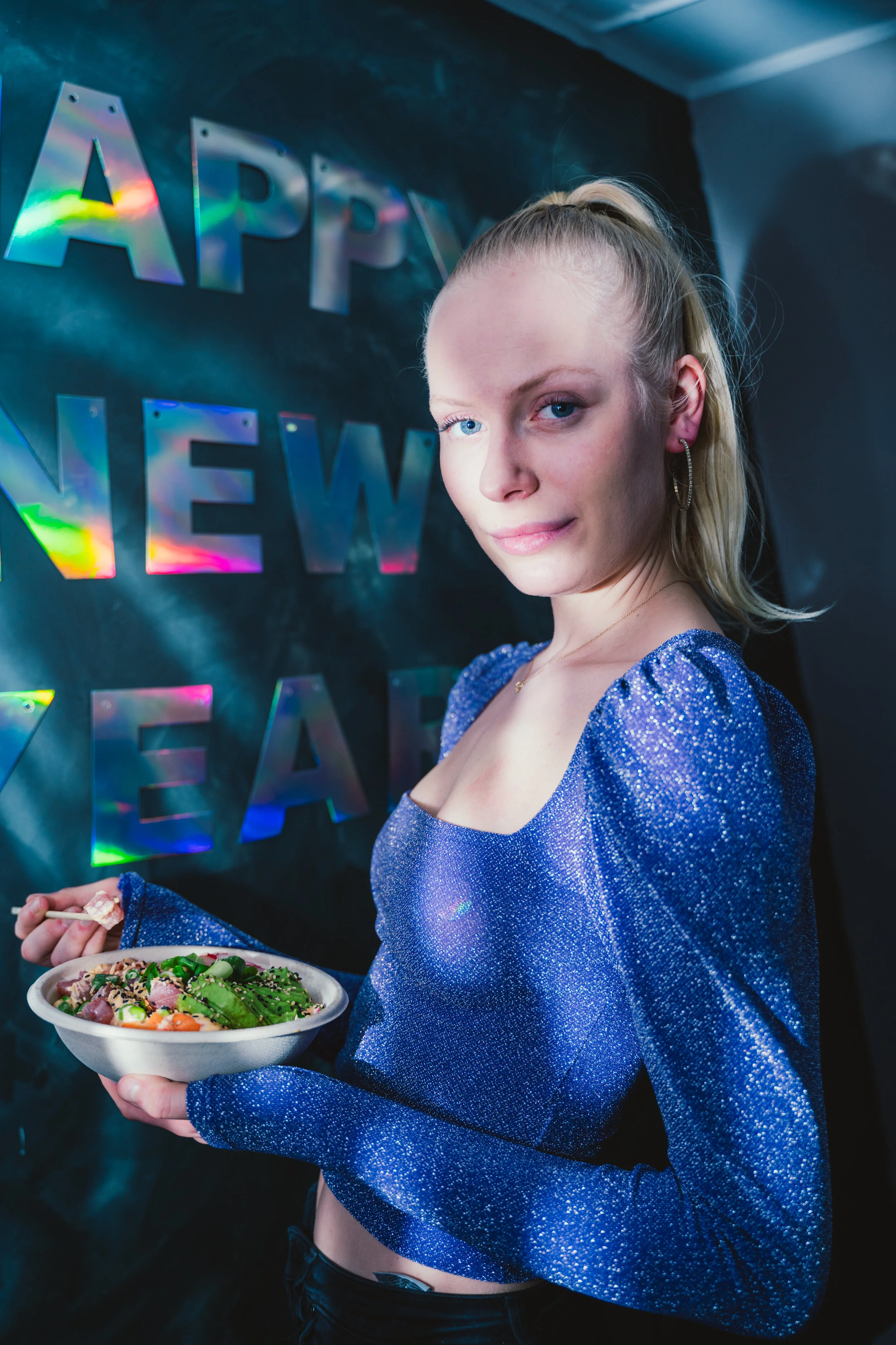 A young woman with blonde hair in a high ponytail, wearing a sparkly blue top, holding a bowl of salad, standing in front of a black wall with iridescent 'Happy New Year' decorations.