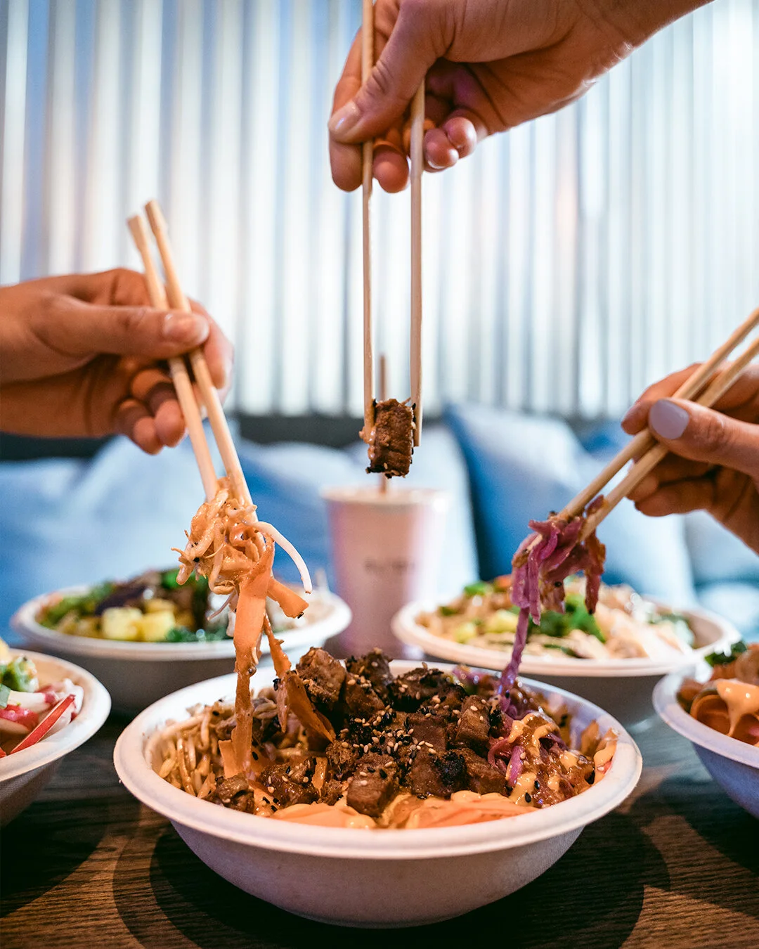 People sharing bowls of Asian noodle dishes with chopsticks, including beef, vegetables, and sauces, in a restaurant setting.