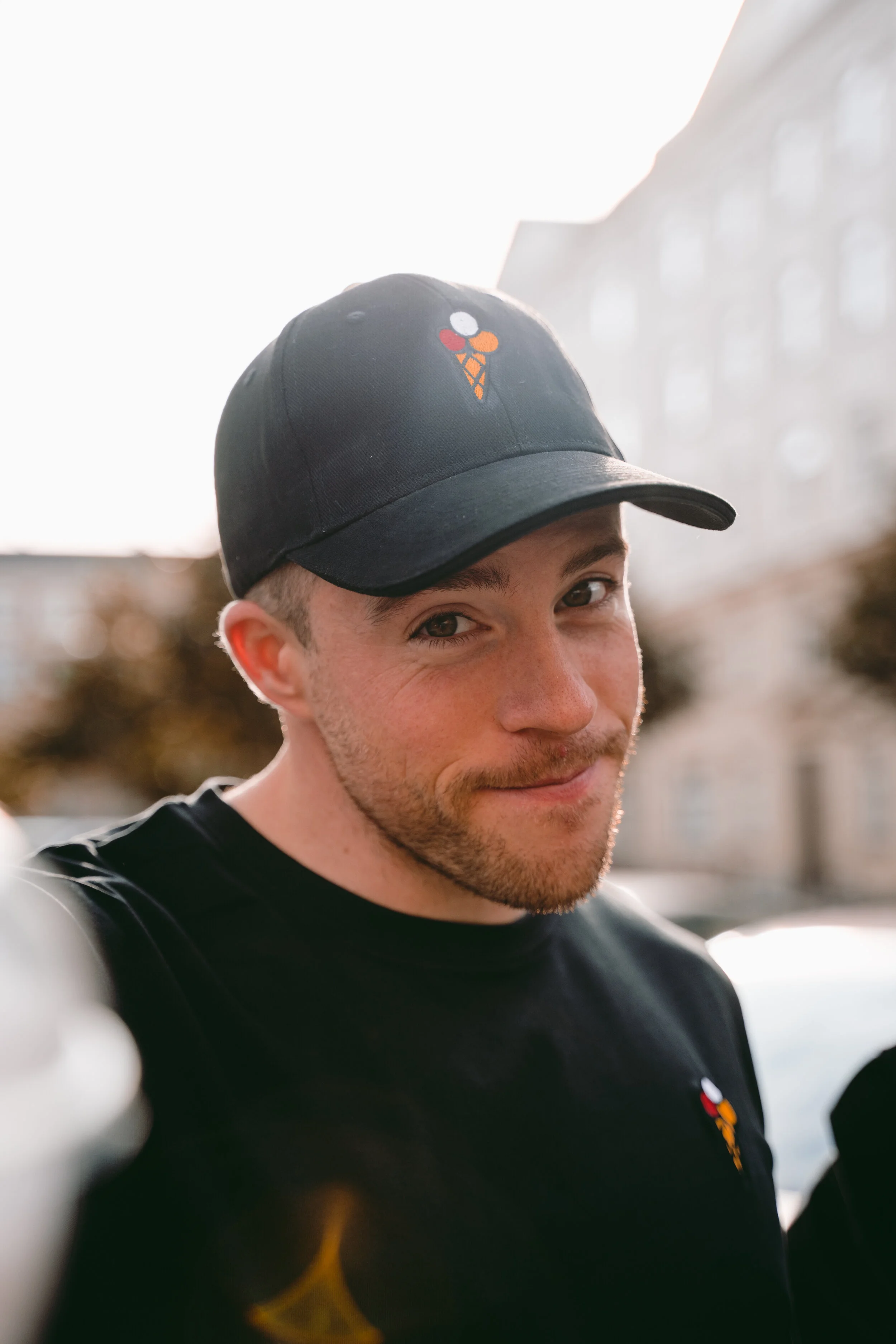 Close-up of a young man wearing a black baseball cap with an ice cream cone logo and a black T-shirt with a small ice cream cone logo, smiling outdoors against a blurred urban background.