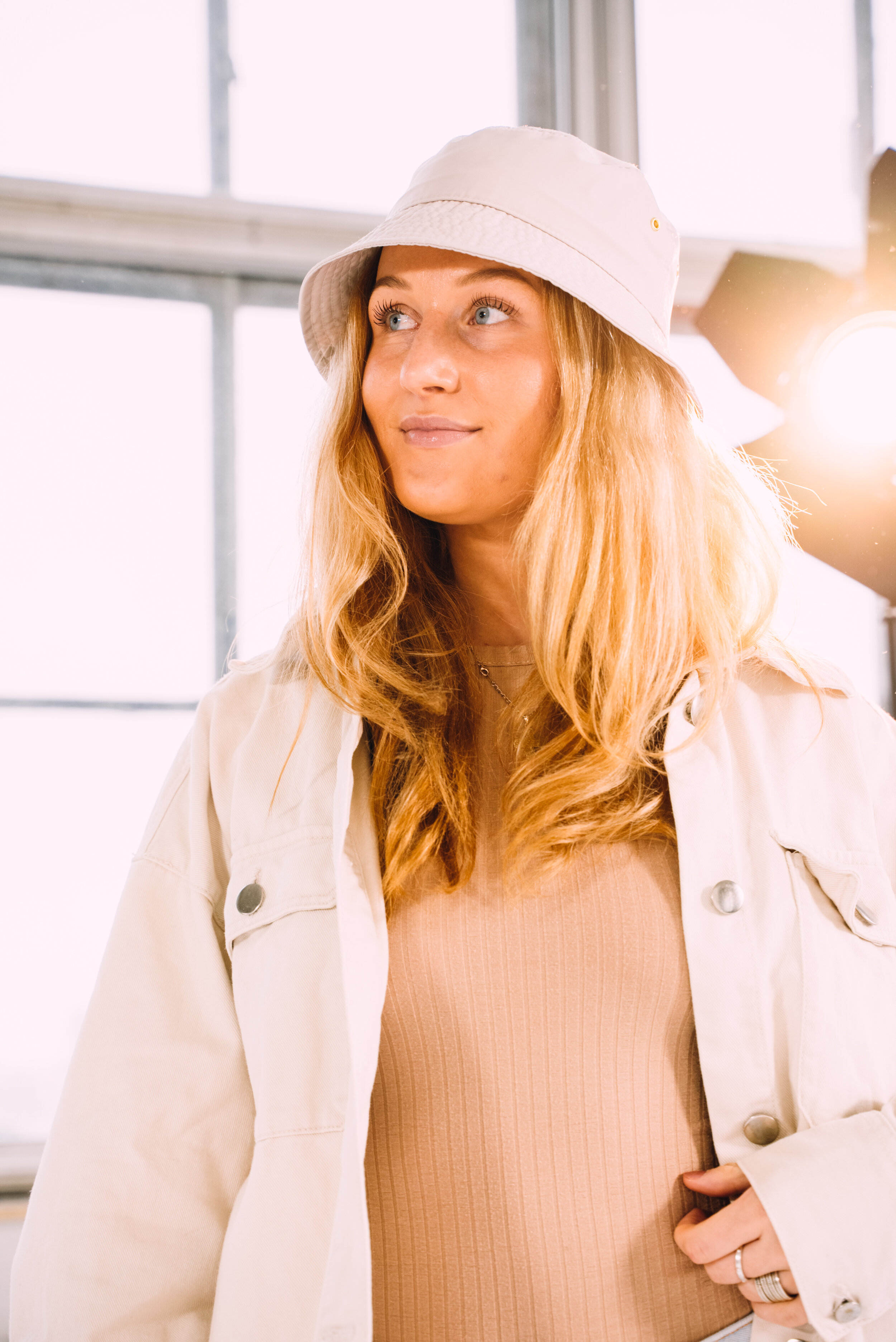 A young woman with long wavy blonde hair wearing a white bucket hat, beige jacket, and beige top stands in a bright indoor setting near a large window, with sunlight shining behind her.