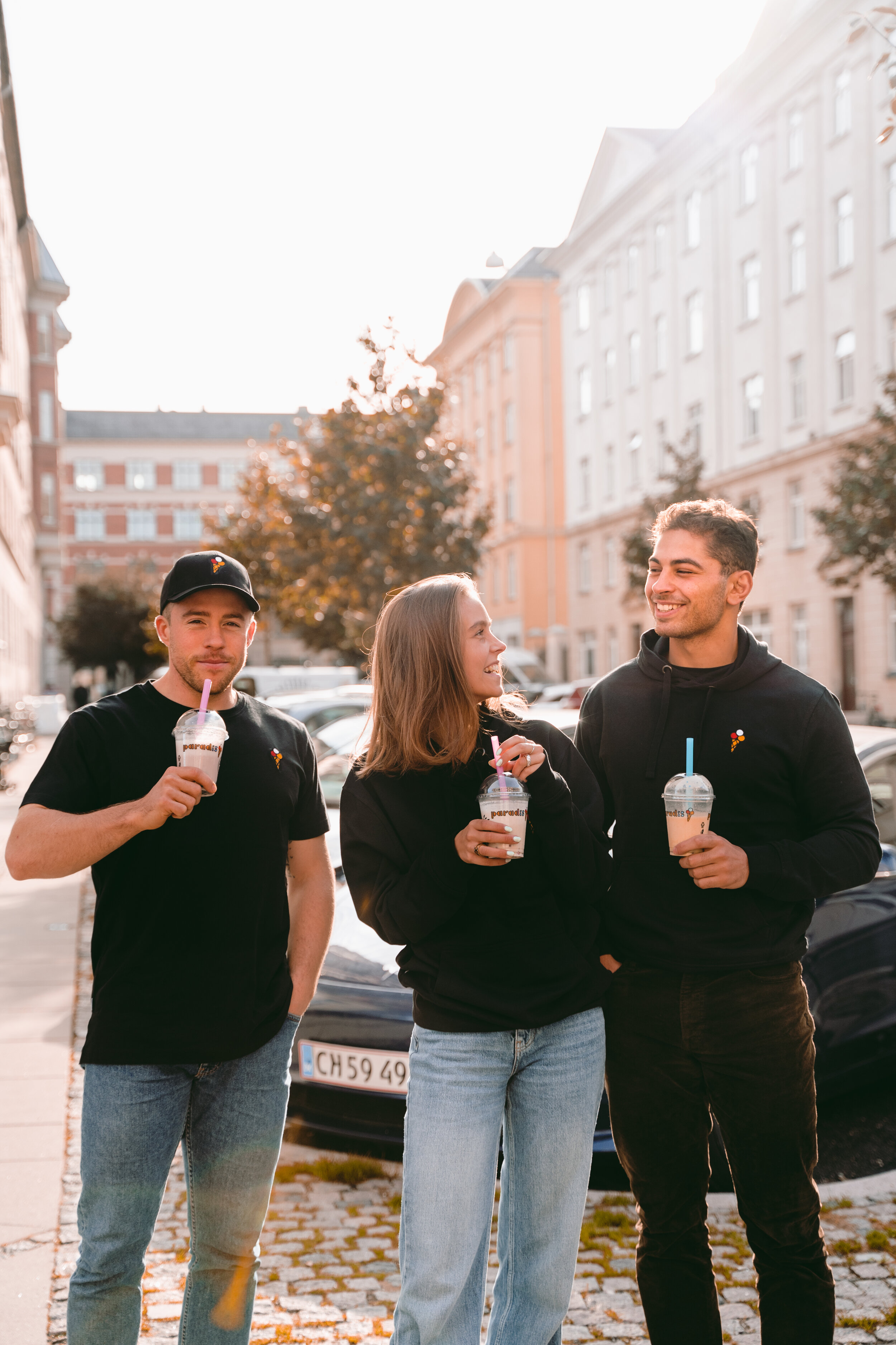 Three young adults standing outside in a city street, smiling and holding drinks, with buildings and parked cars in the background.