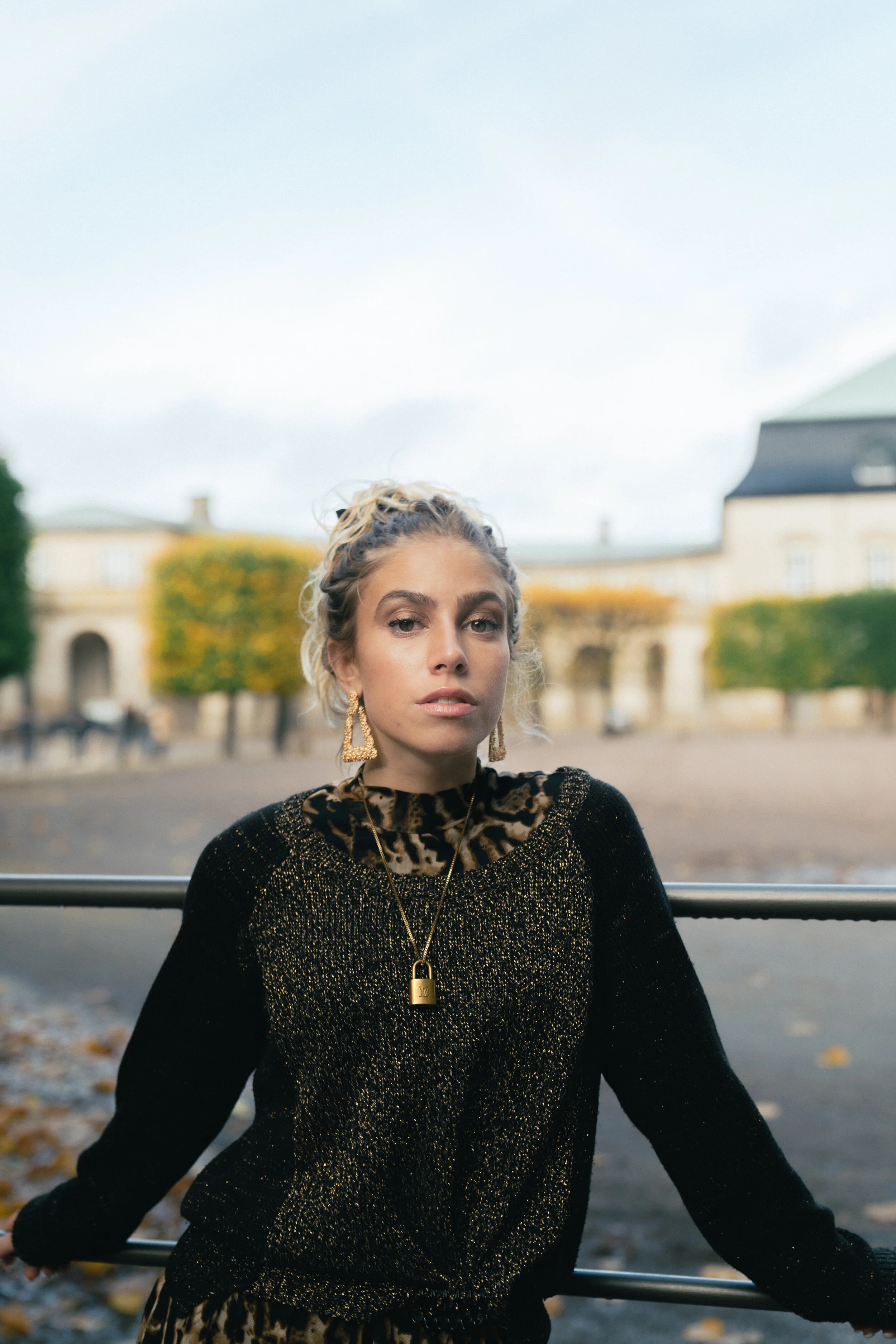 A young woman outdoors during daytime, wearing a black and gold sweater, leopard print turtleneck, and gold jewelry, standing in front of a historic European-style building with trees.