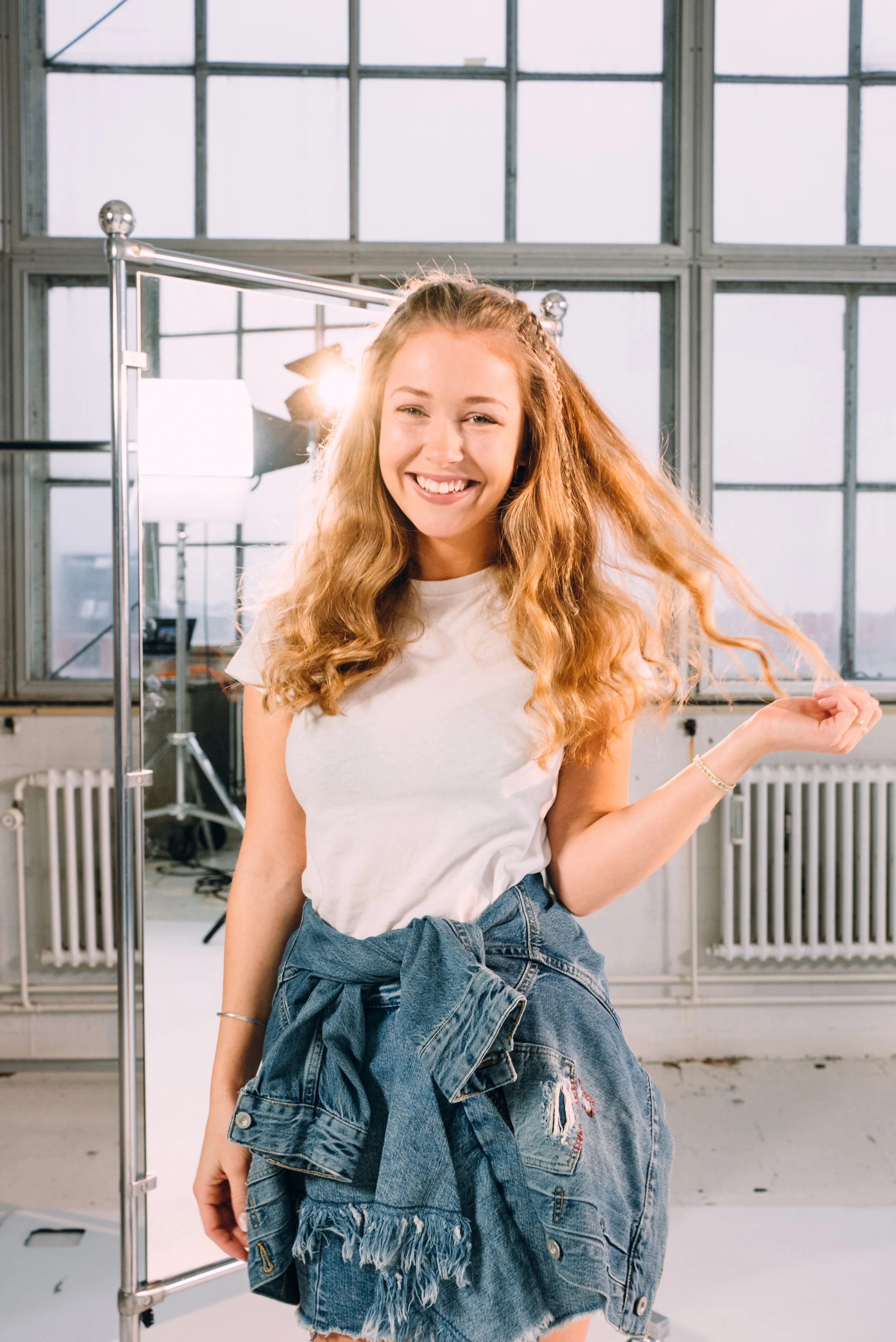 Young woman with curly red hair in a studio with large windows, smiling and posing with one hand raised.