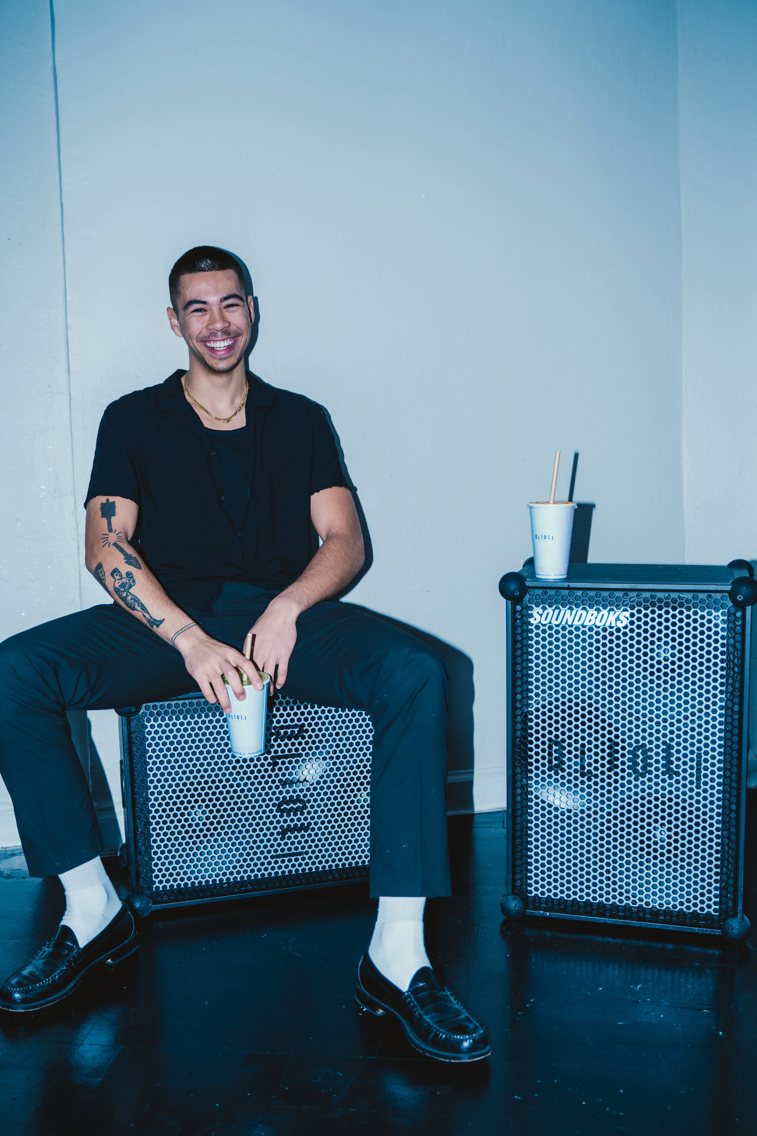 A young man with tattoos, dressed in black, sitting on a speaker with a smile, holding a cup, with another cup on an amplifier beside him, in a minimalistic room.